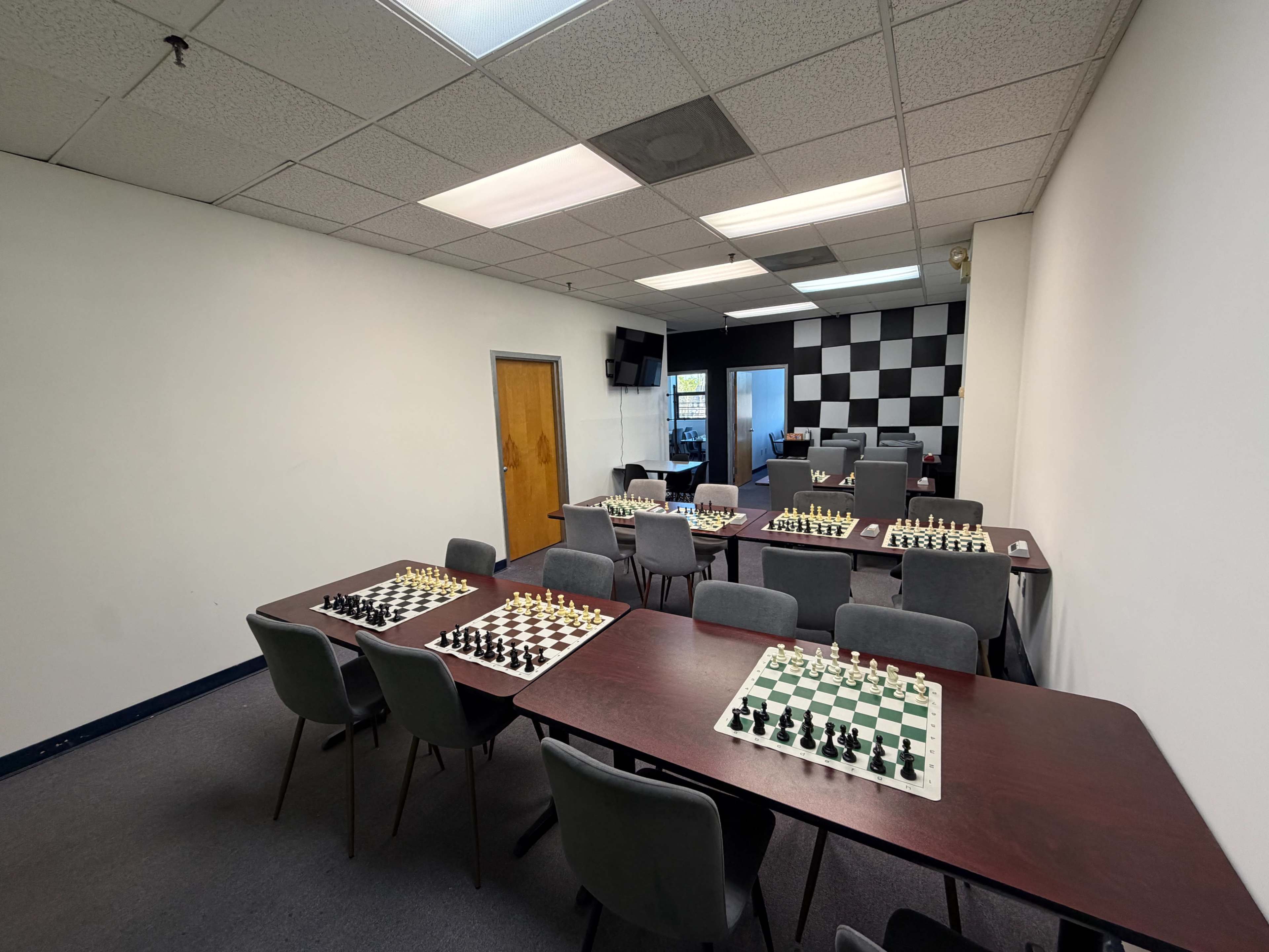 The image shows a room set up for chess, featuring several tables with chessboards arranged for gameplay and a checkered wall in the background.