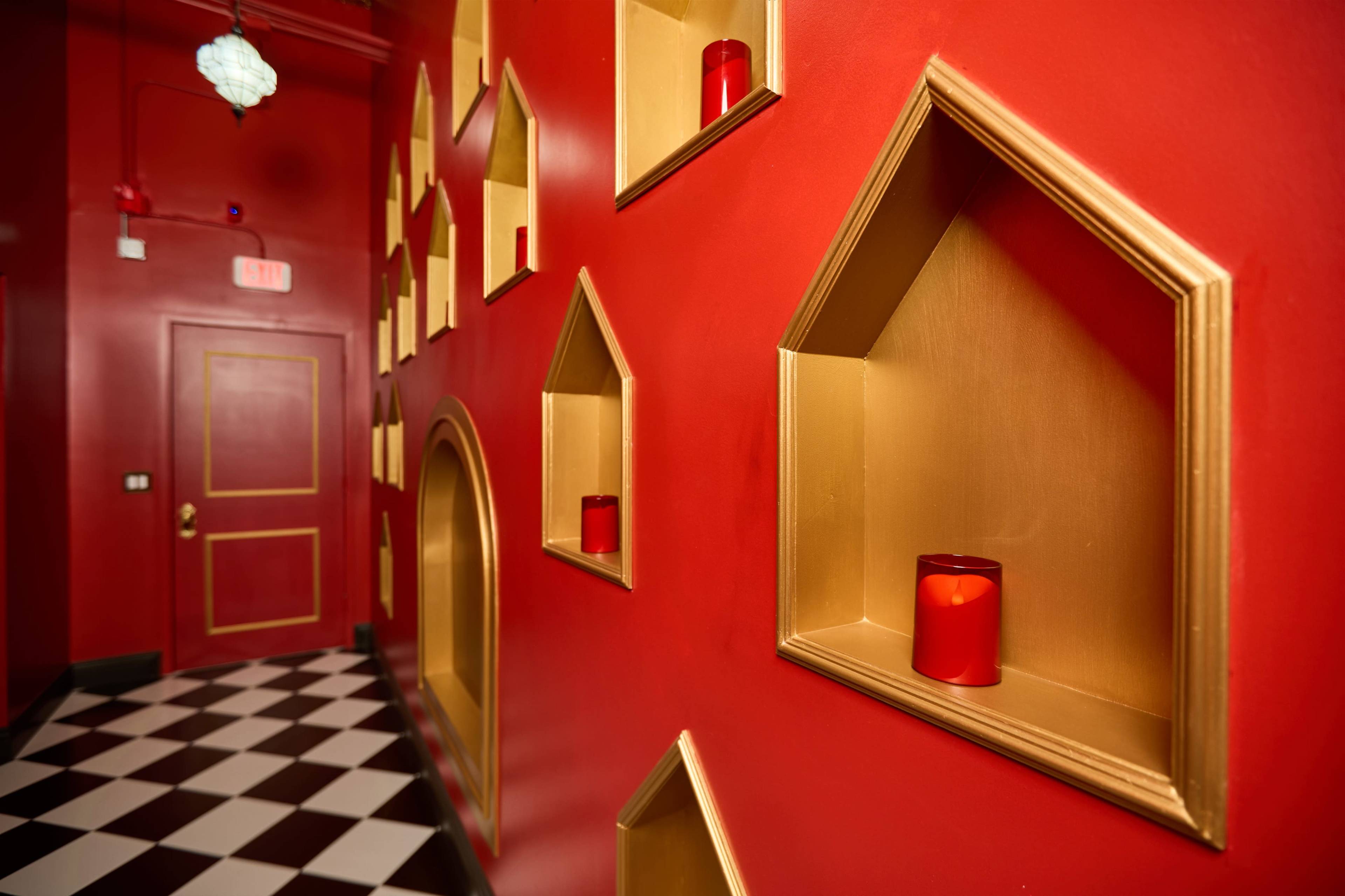 The image displays a hallway with a vibrant red wall featuring golden, shaped alcoves adorned with candles, and a checkered black and white floor.