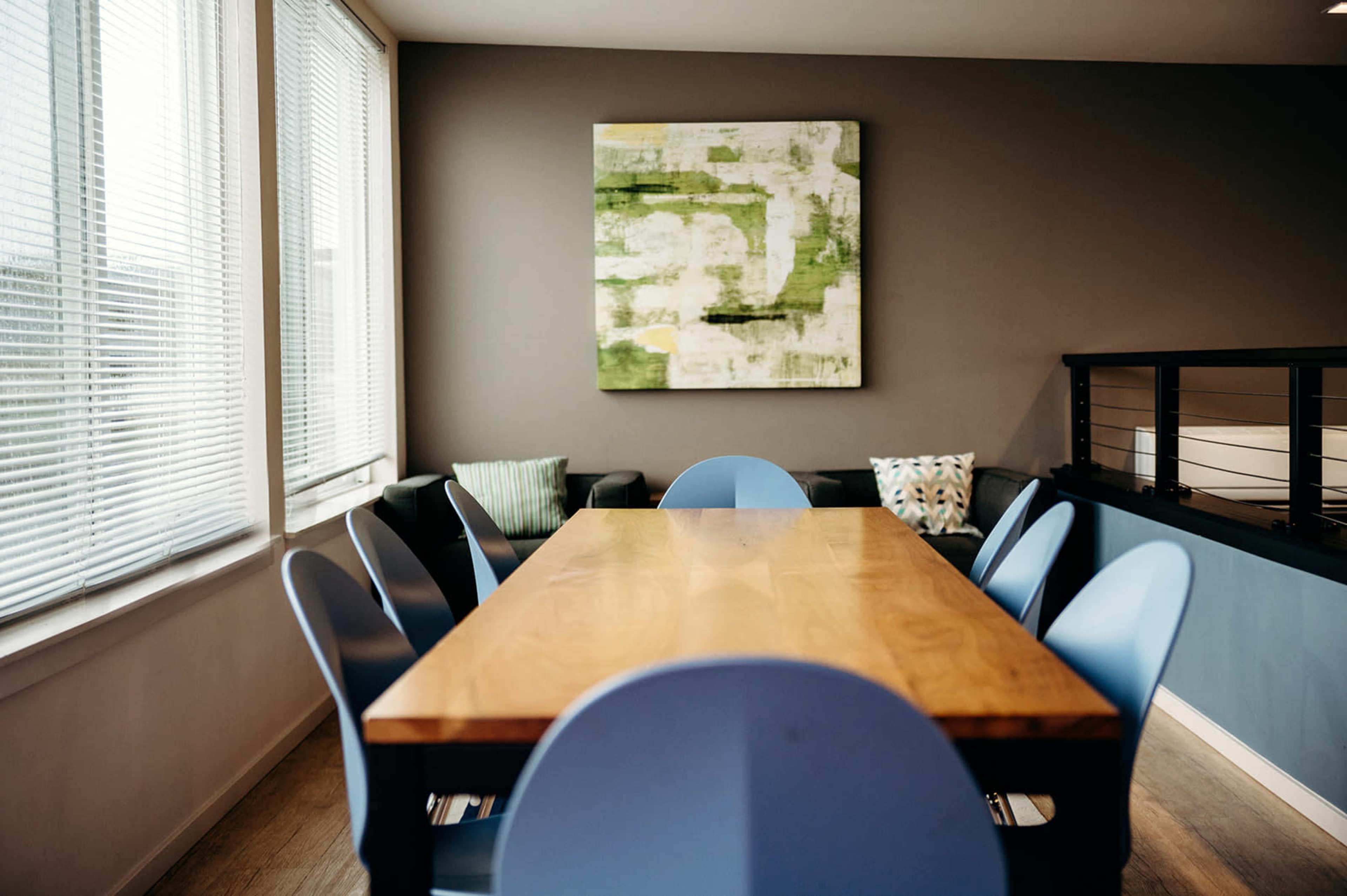 A rectangular wooden table with blue chairs is set in a well-lit room featuring large windows and a green and white abstract painting on the wall.
