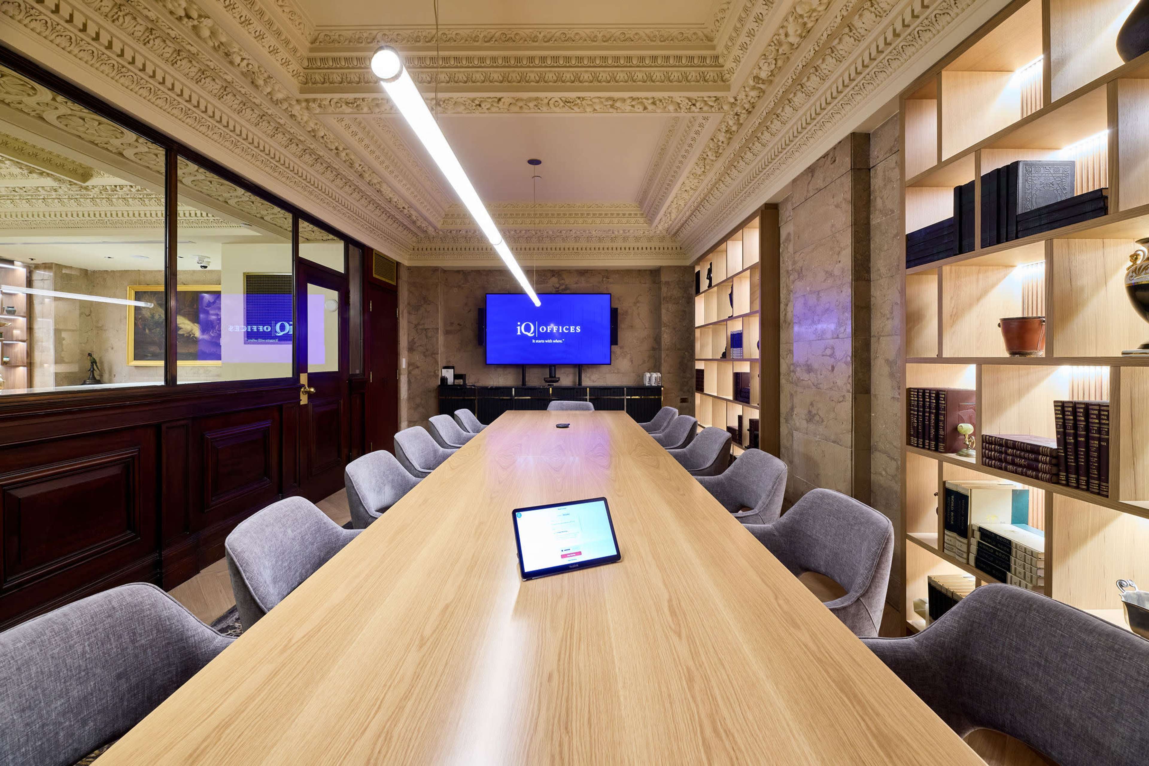 A long wooden conference table is set in a well-lit meeting room, with chairs arranged around it and a large screen displaying a logo on the wall.
