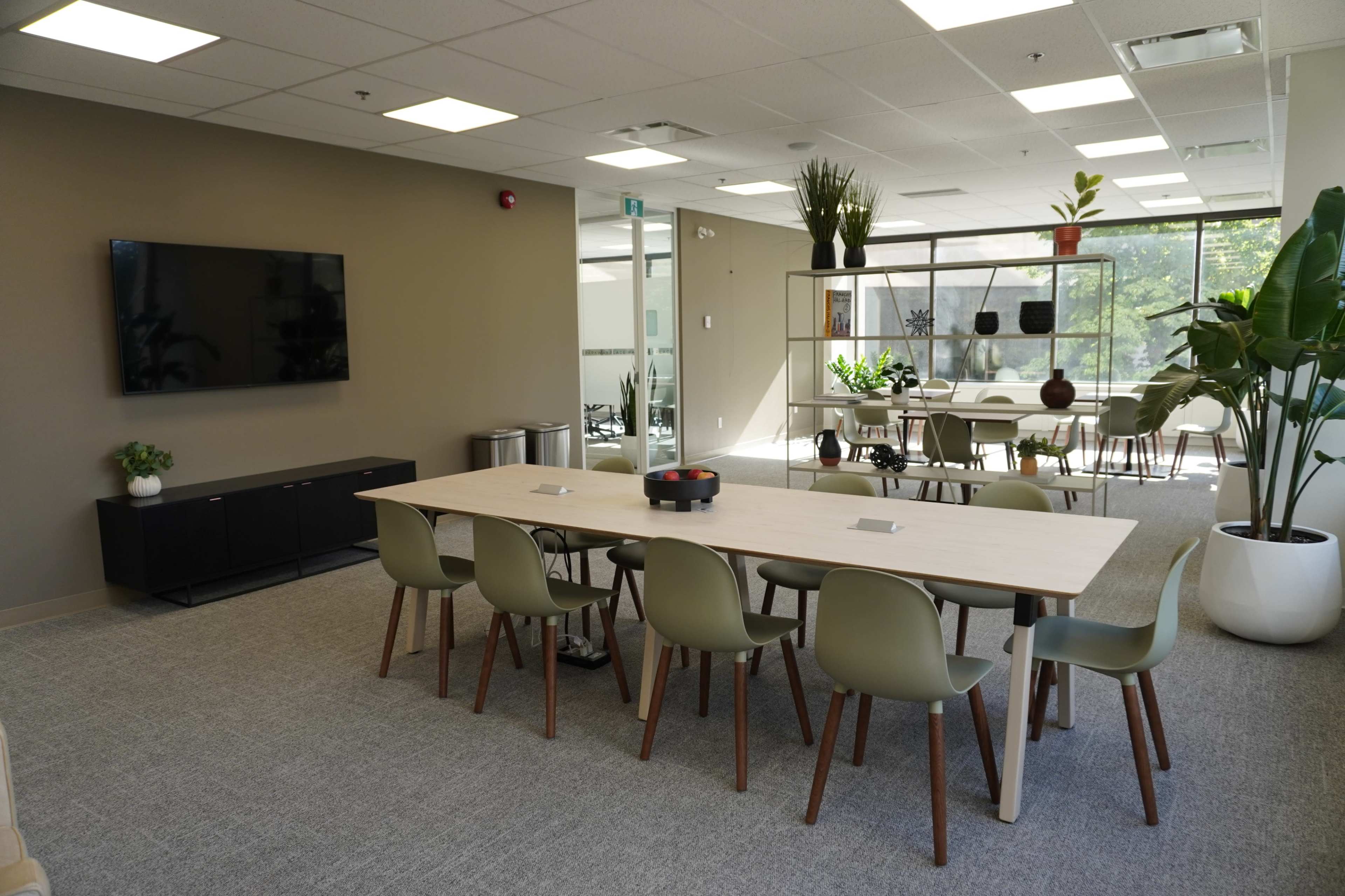 The image shows a modern conference room with a long wooden table surrounded by green chairs, a flat-screen TV mounted on the wall, and large windows letting in natural light.