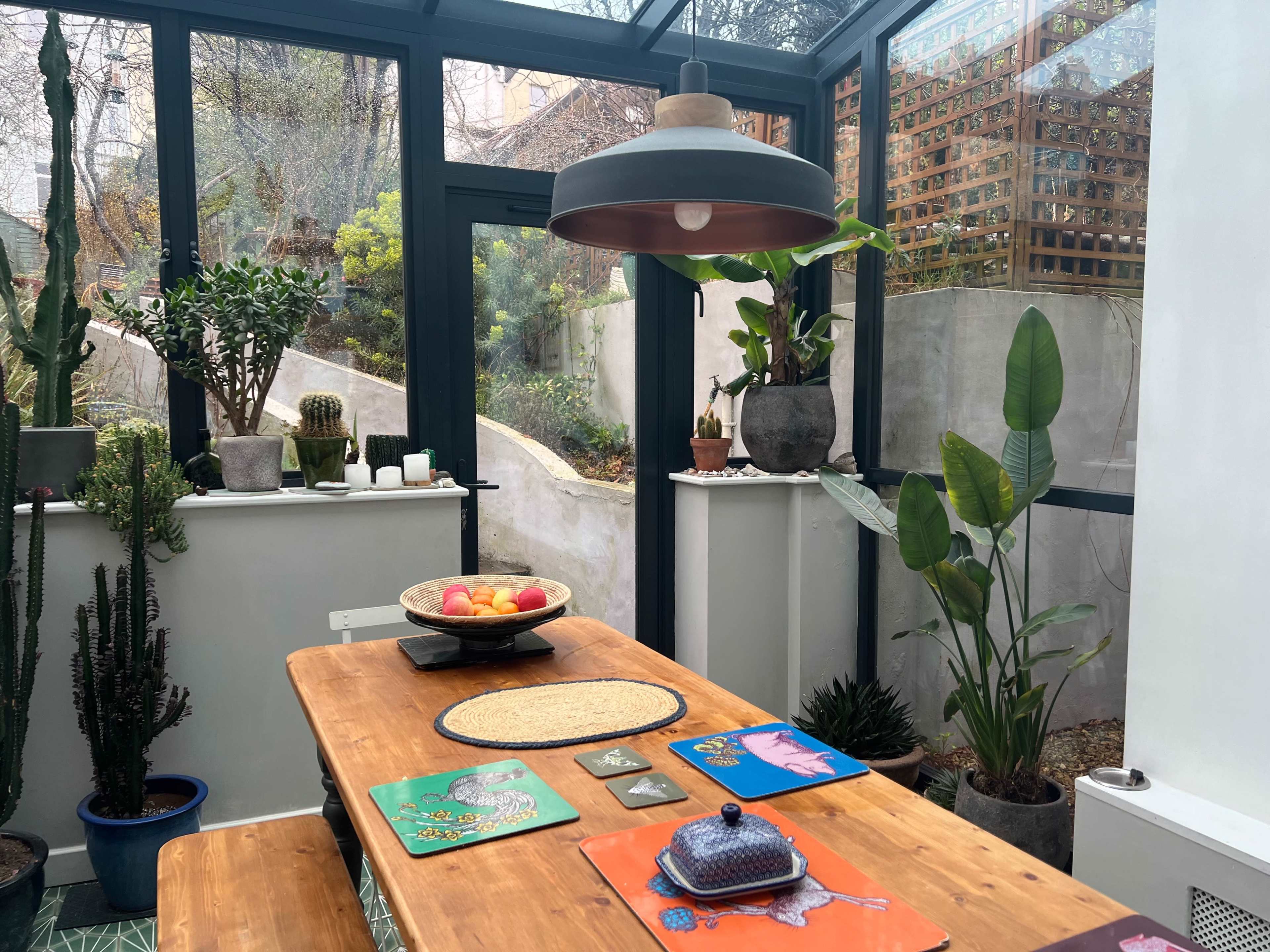 A wooden dining table is set in a sunlit room filled with plants, featuring decorative table mats and a bowl of apples at the center.