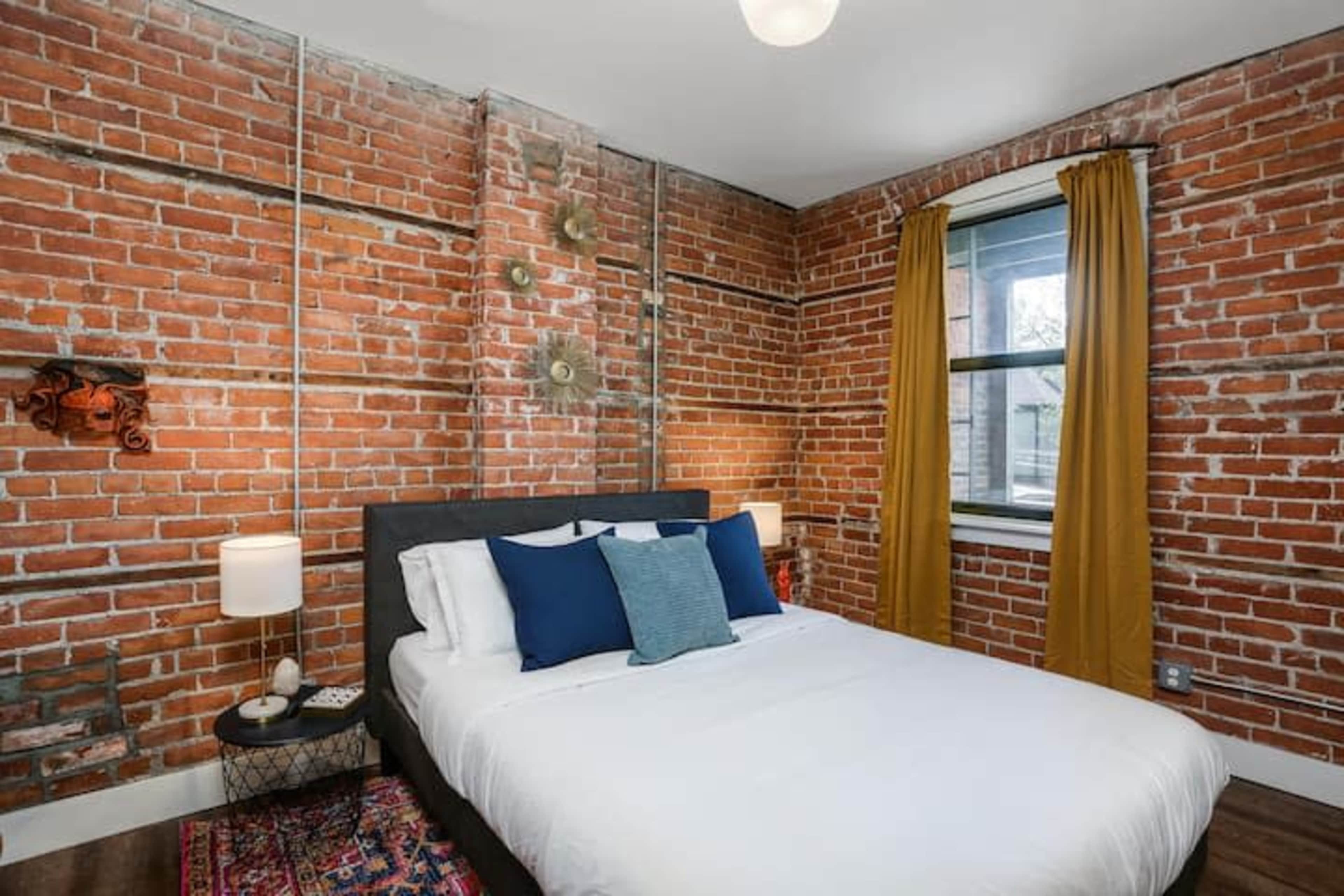 A bedroom with exposed brick walls, a bed with white linens and blue accent pillows, and a window draped with yellow curtains.