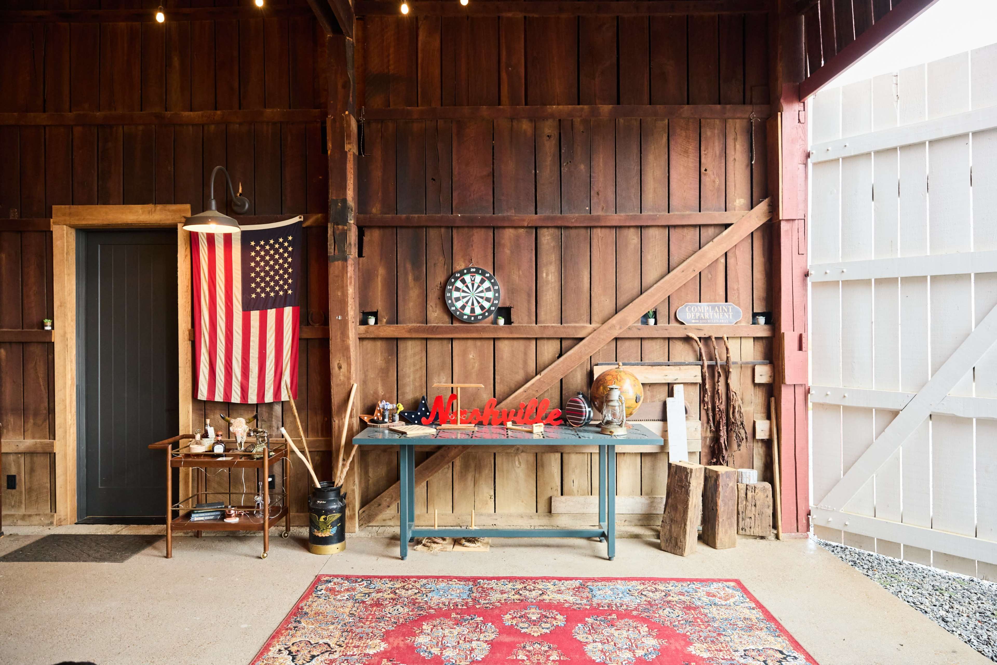 The image shows a rustic barn interior featuring a wooden wall, an American flag, a dartboard, and a table with various decorative items and a rug on the floor.