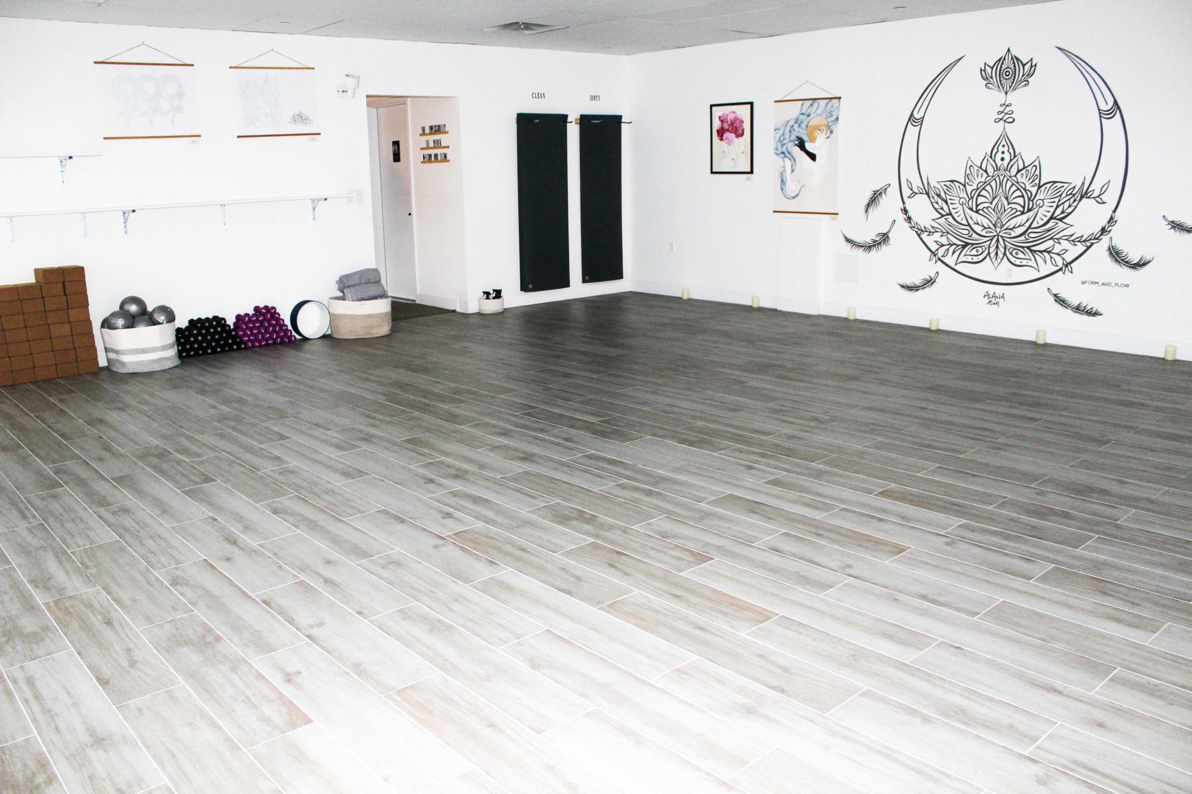 An empty studio space with wooden floors, yoga mats stacked on the left, and wall decorations including artwork and a mural.