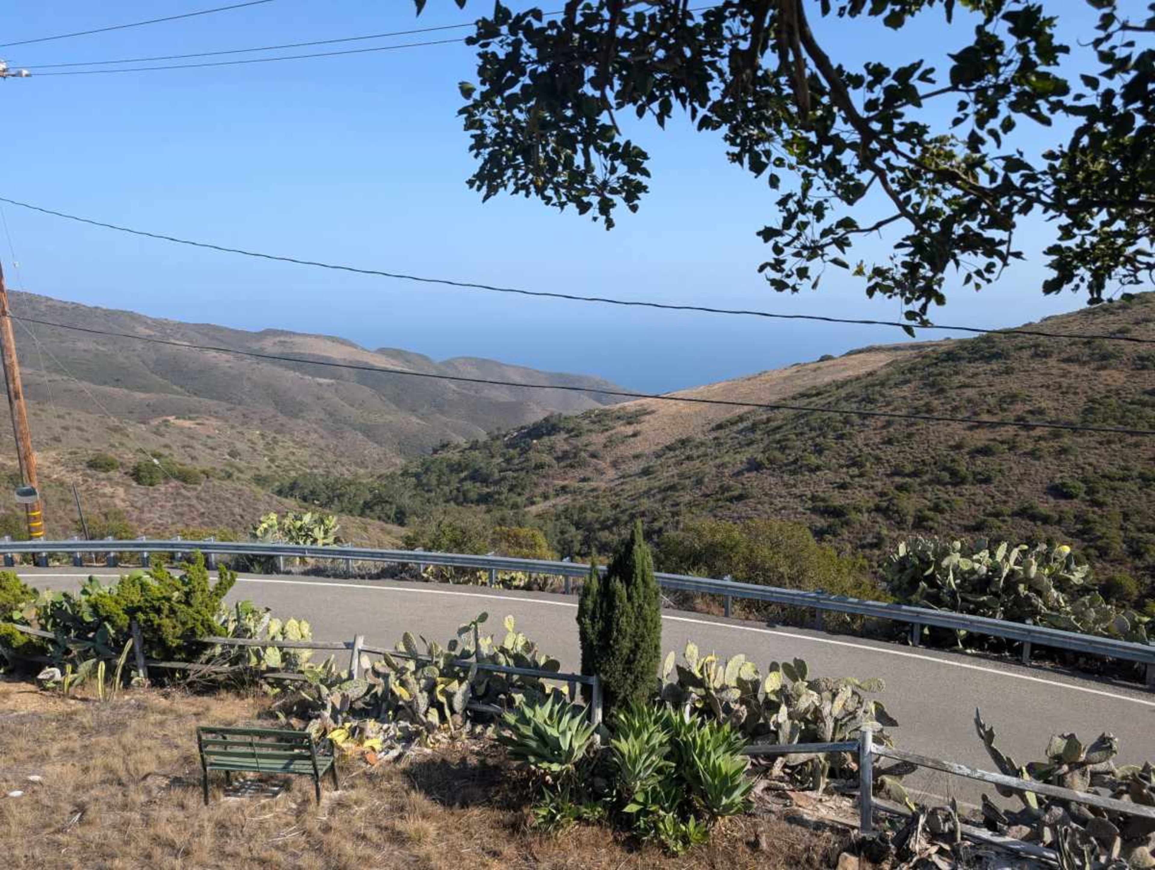 The image shows a winding road cutting through hilly terrain with sparse vegetation and a view of the ocean in the distance.
