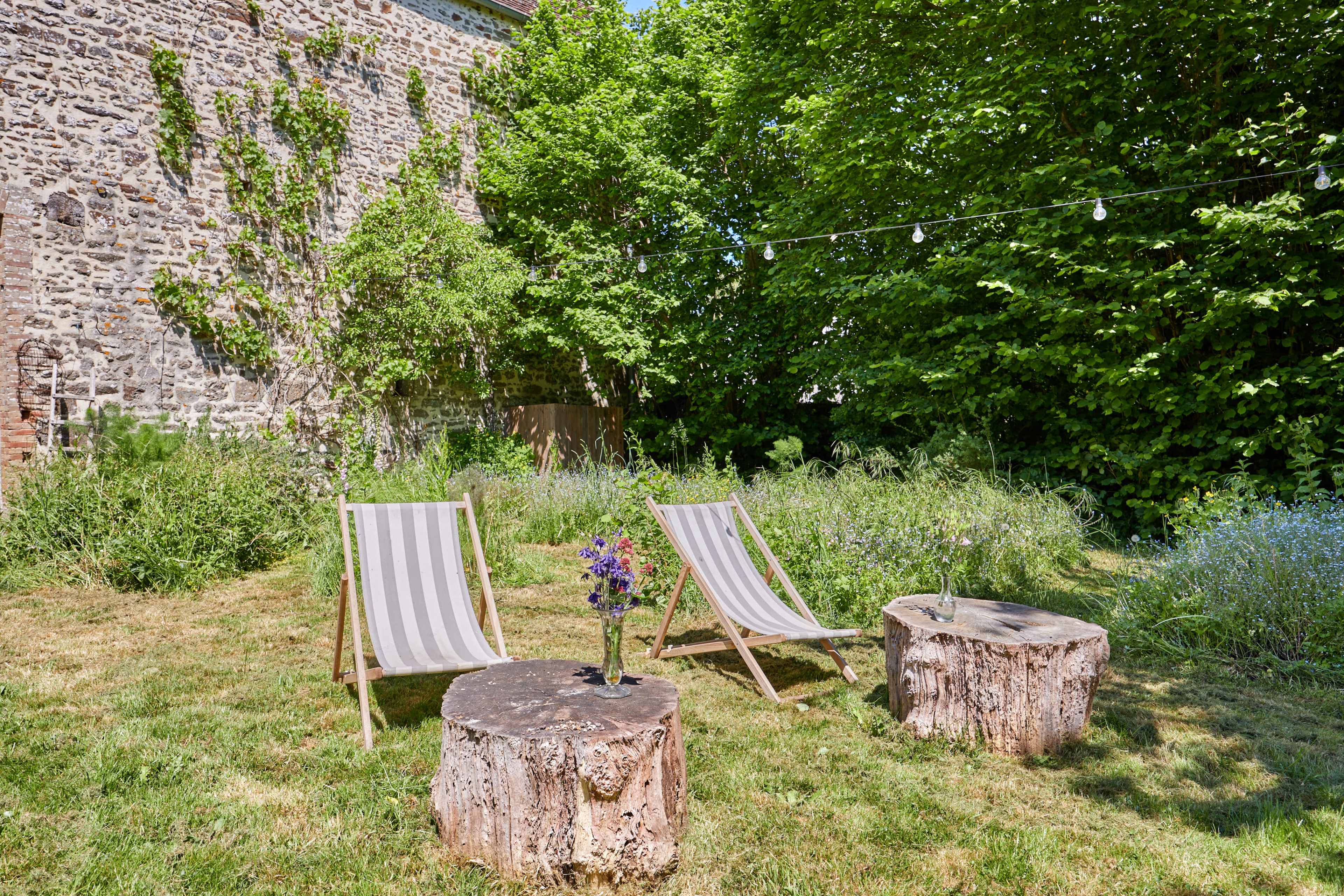 The image shows two striped lounge chairs placed on a grassy area surrounded by greenery, with two wooden stump tables and a small bouquet of flowers on one table.