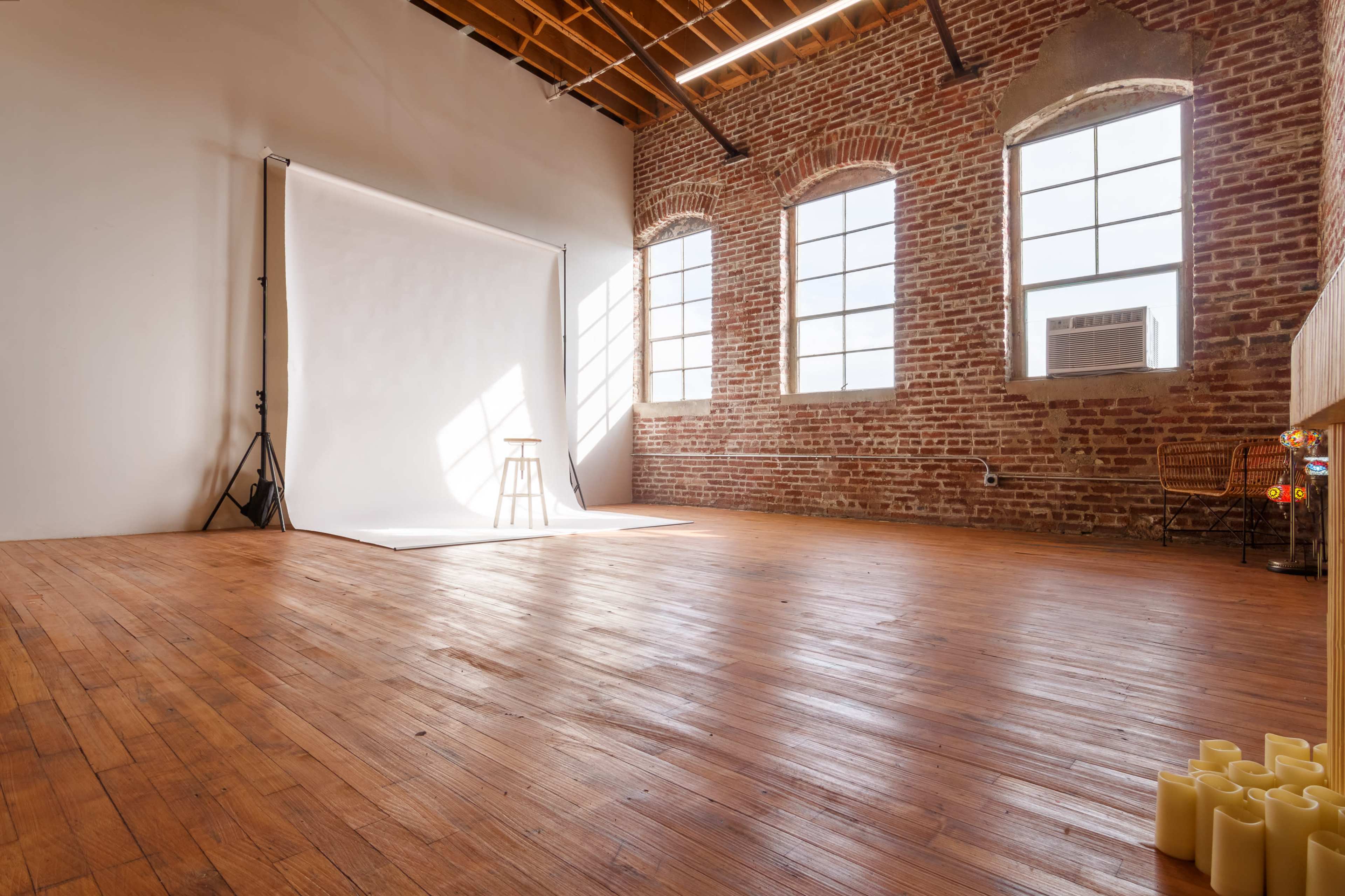 A spacious studio with brick walls, large windows, and wooden flooring, featuring a white backdrop and a stool in the center.