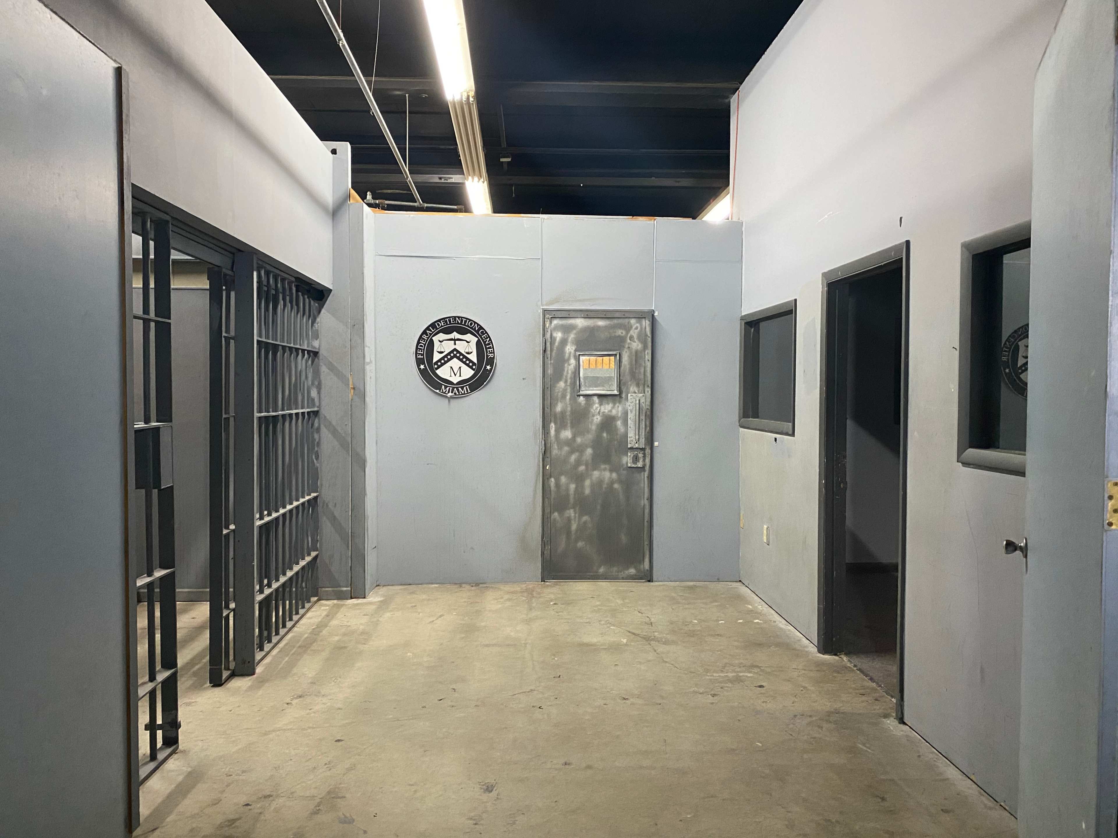 The image shows an interior space resembling a police station or jail, featuring a barred cell on the left, a solid door at the far end, and several plain walls with windows and a wall emblem.