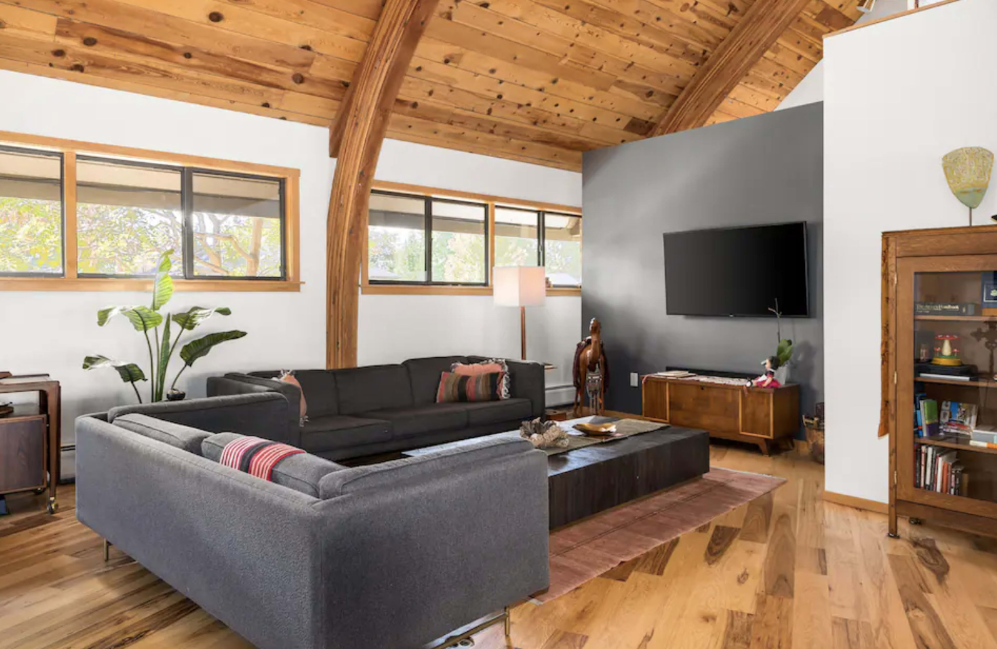 A modern living room featuring a dark sectional sofa, a central coffee table, wooden ceilings, and a mounted television on a gray wall.