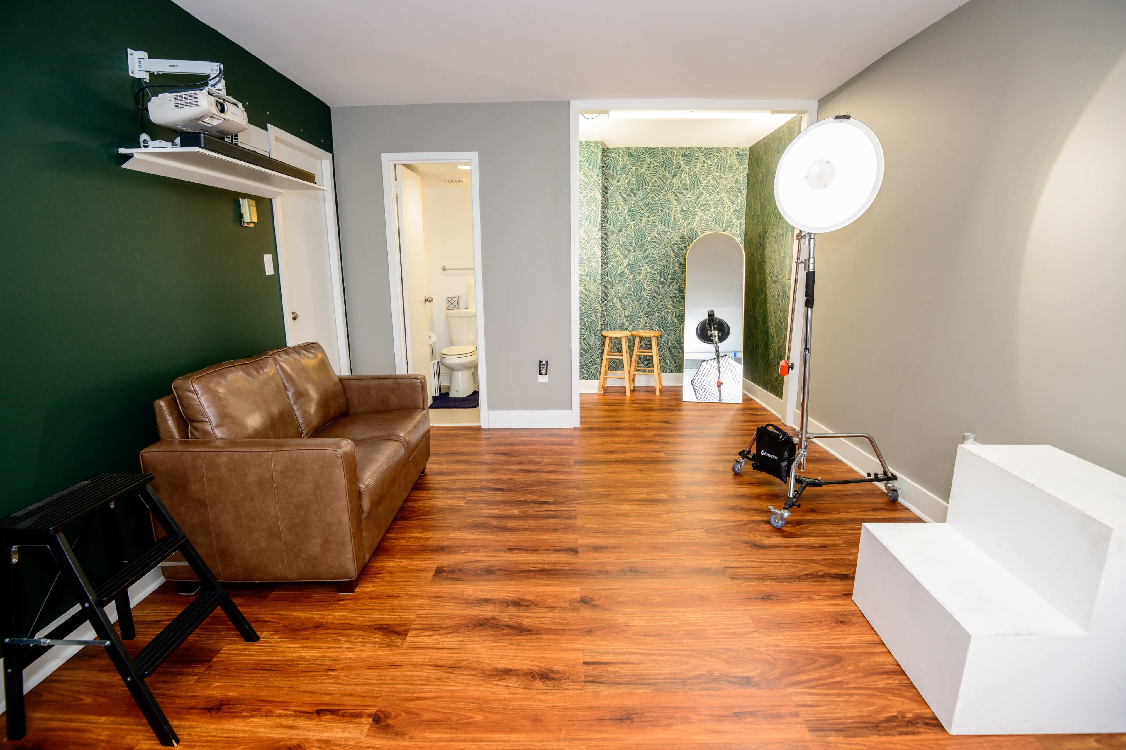 An indoor space featuring a brown sofa, a projector on a shelf, a green accent wall, wooden flooring, and a small staircase leading to a well-lit area with a mirror and stools.