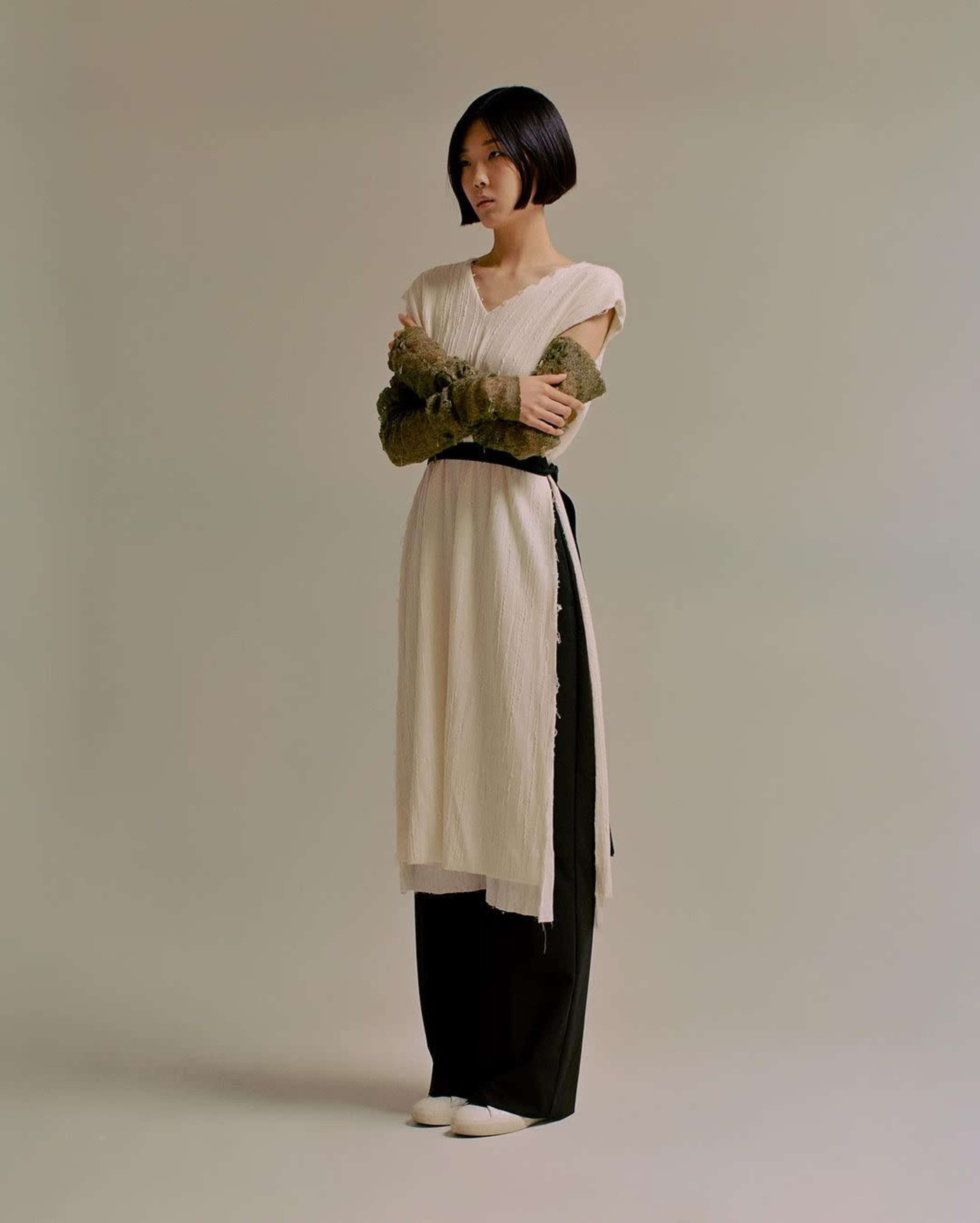 A woman stands with her arms crossed, wearing a long, layered outfit consisting of a light-colored dress over dark trousers.