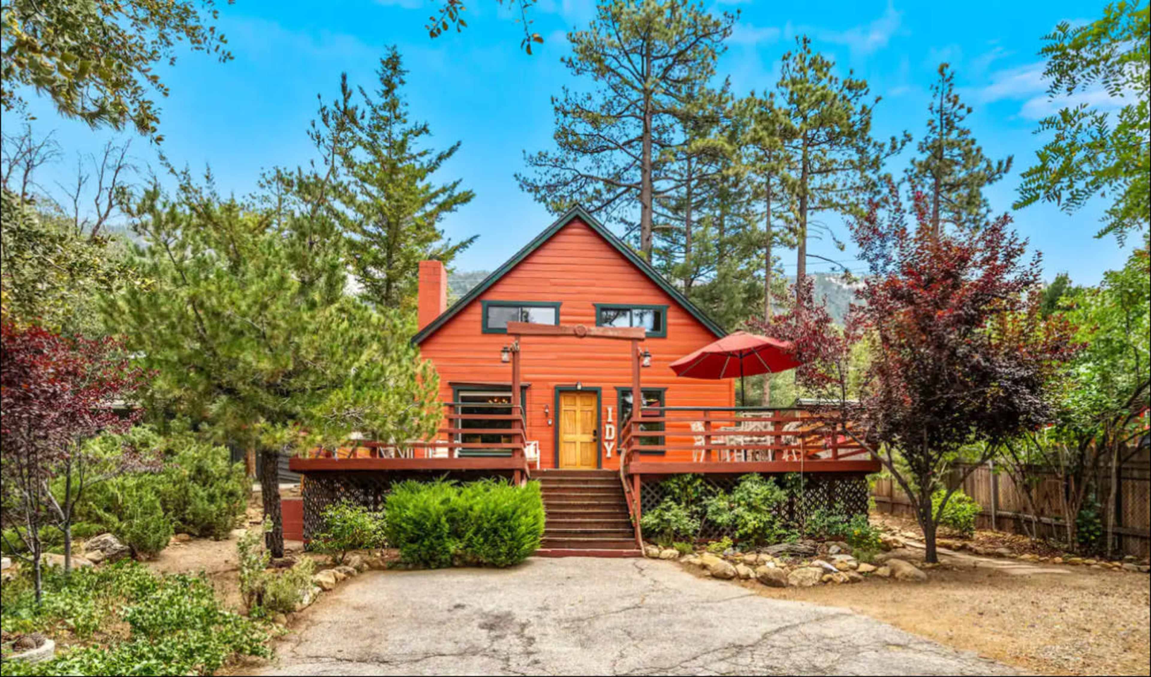 A red-brick house with an A-frame roof is surrounded by trees and features a front deck with an umbrella.