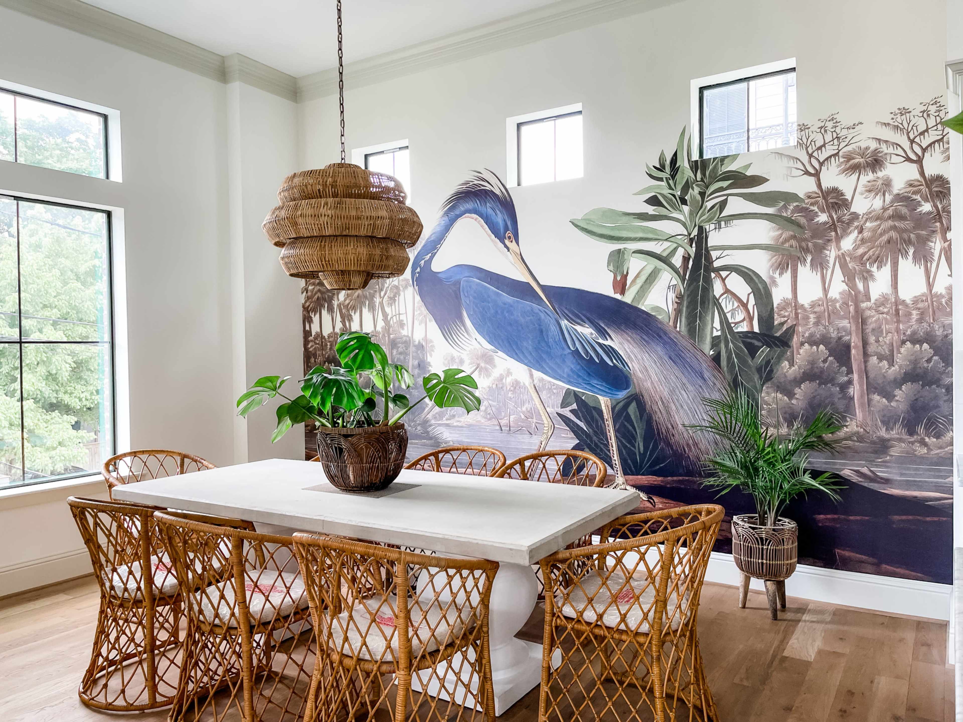 A dining room features a large mural of a blue heron and a table surrounded by wicker chairs with a potted plant in the center.