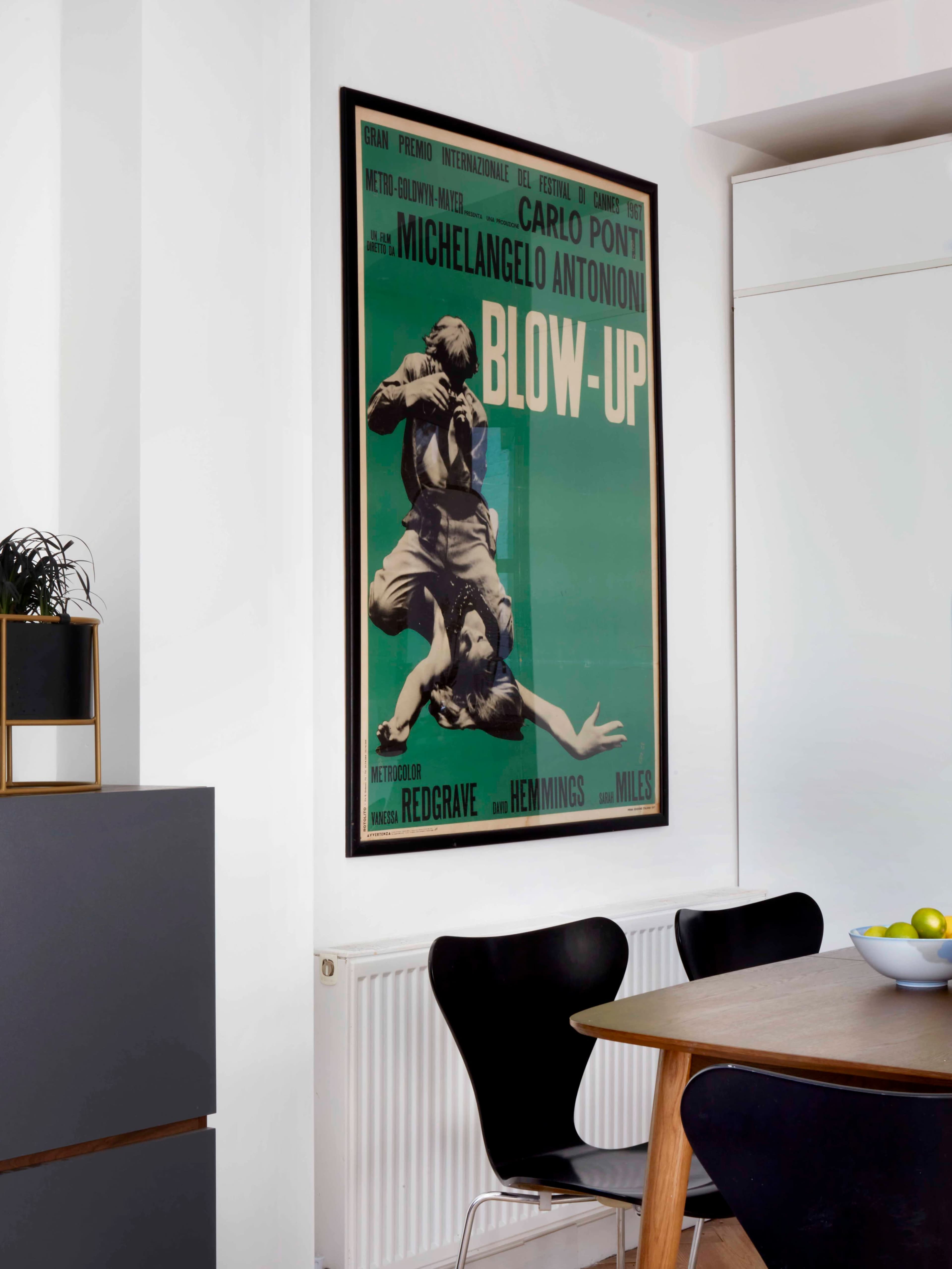 A vintage movie poster for "Blow-Up" by Michelangelo Antonioni hangs on a white wall above a wooden dining table with black chairs.