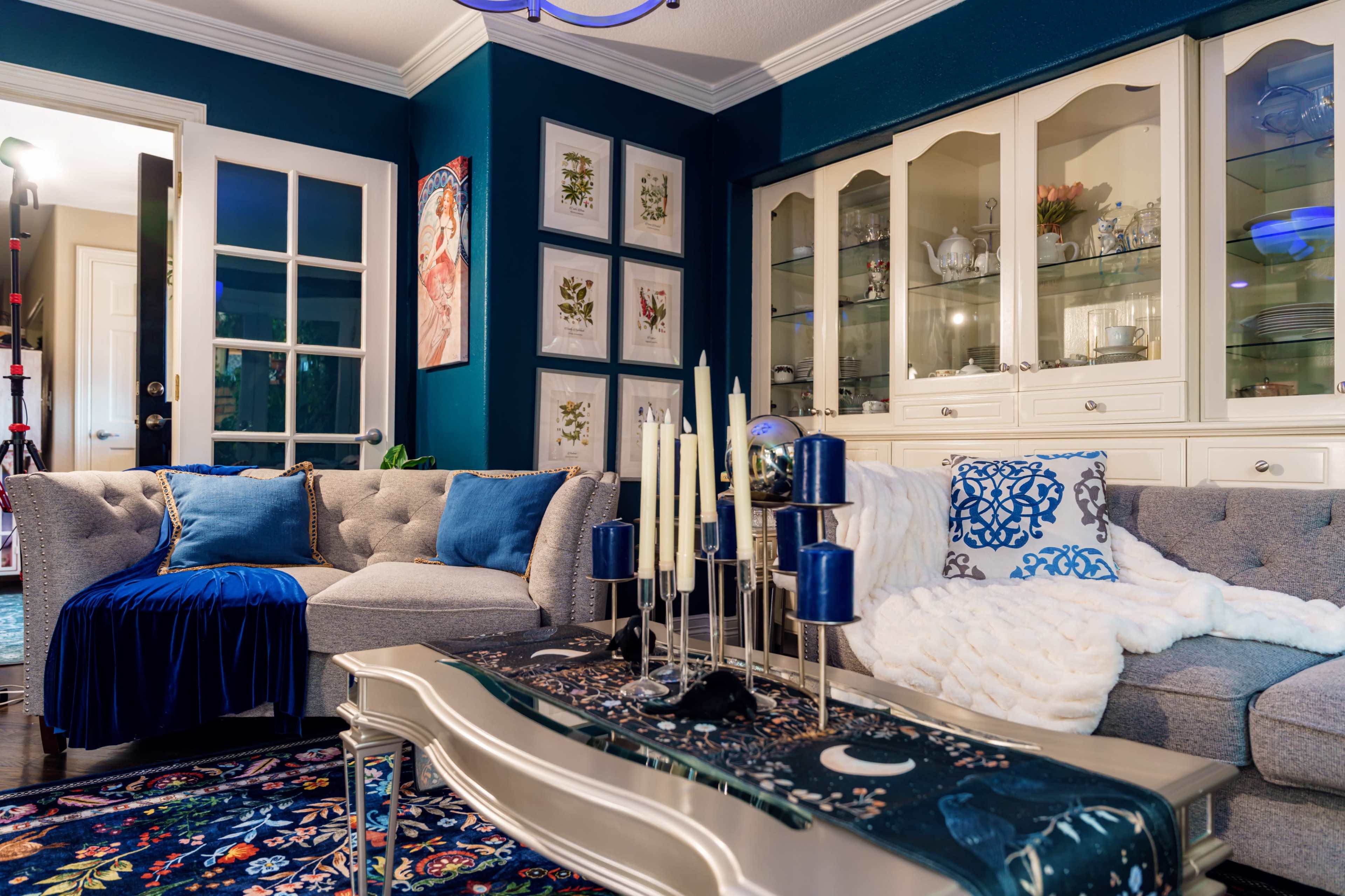 The image features a cozy living room with a gray sectional sofa, decorative pillows, and a glass coffee table centered on an ornate rug, surrounded by blue walls and a display cabinet filled with dishware.