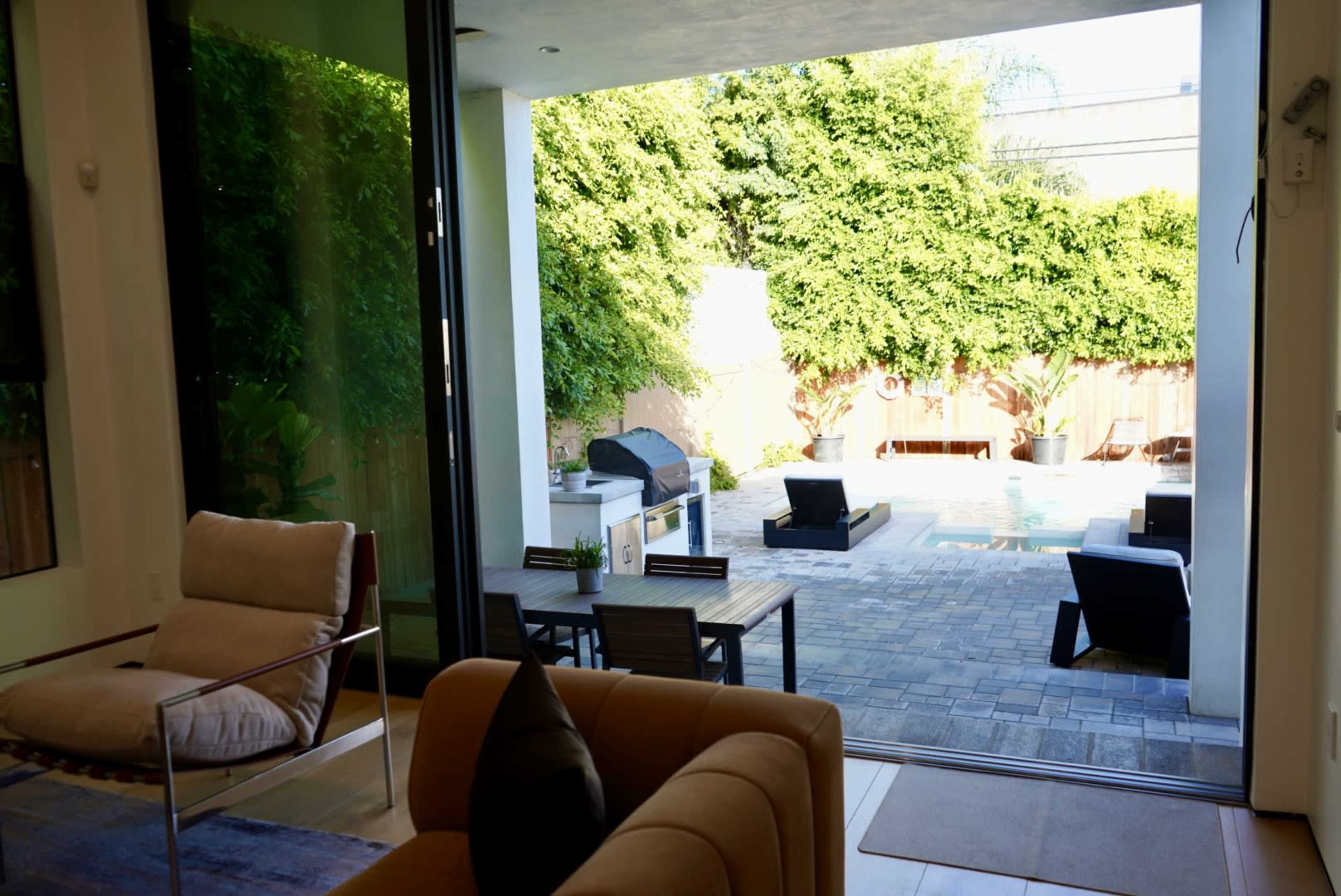 The image shows a modern indoor space with large glass doors opening to a backyard featuring a swimming pool, patio furniture, and a barbecue area surrounded by greenery.