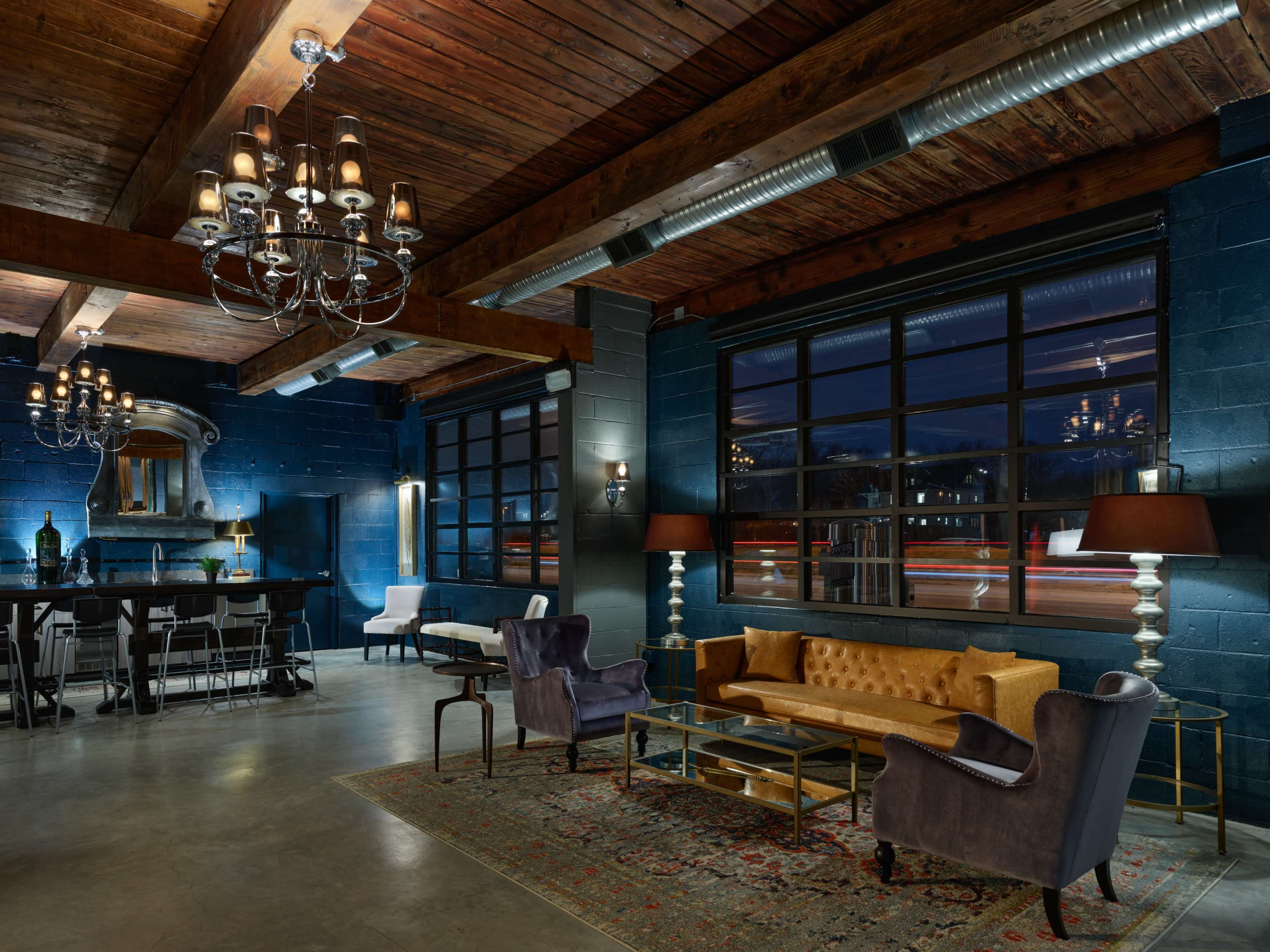 The image shows a spacious, modern lounge with wooden beams, elegant chandeliers, and stylish furniture, highlighted by large windows that reveal a nighttime cityscape.