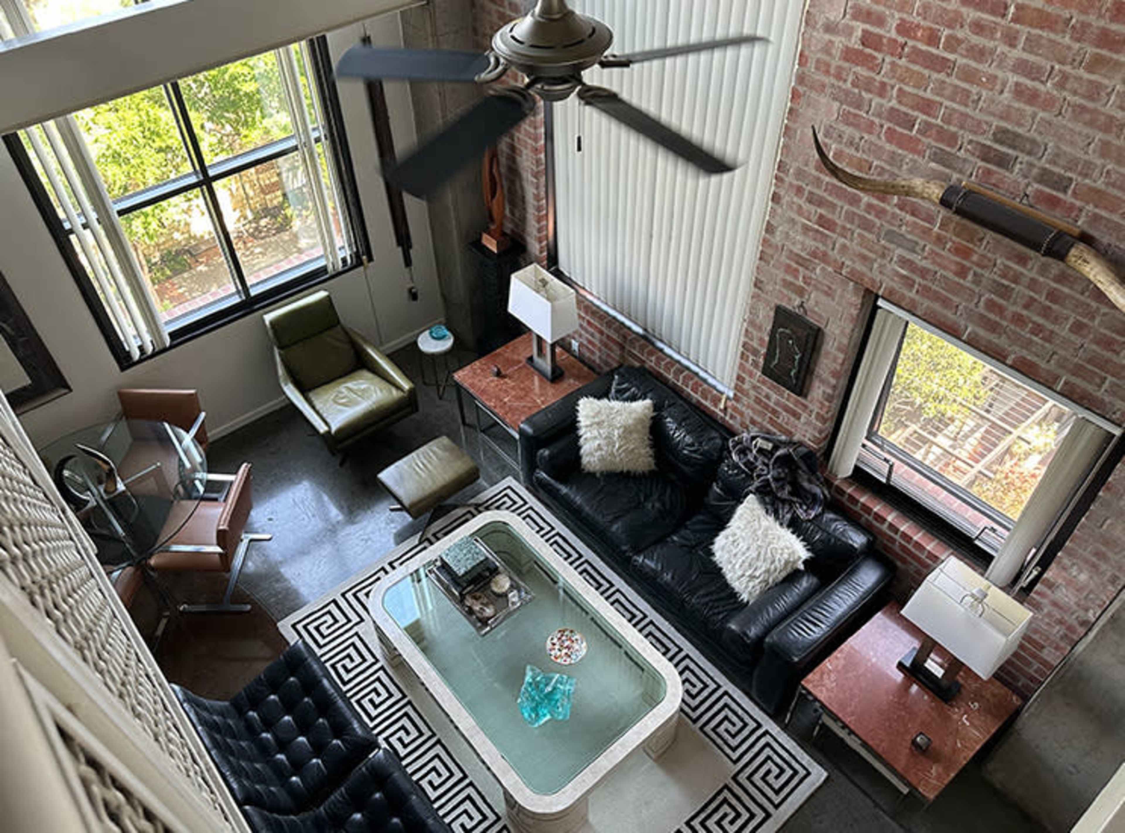The image shows a modern living space with a large black leather sofa, a glass coffee table, and two armchairs, all surrounded by brick walls and large windows.