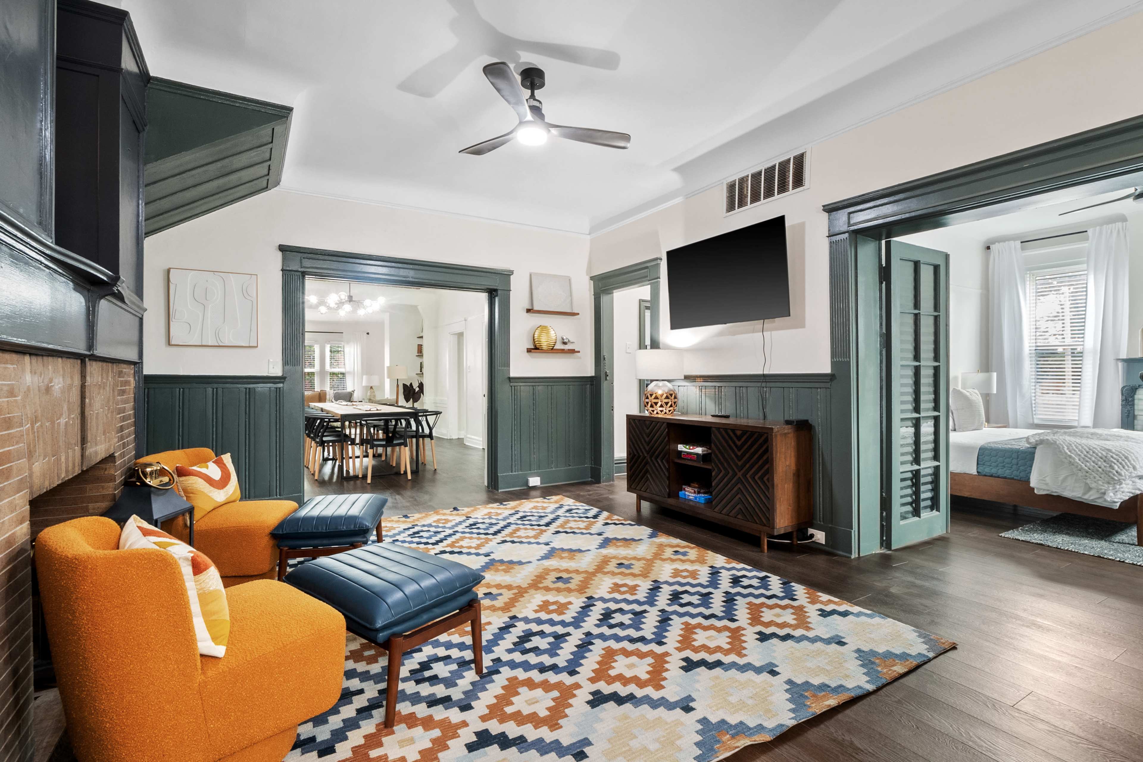 A well-furnished living room featuring two orange chairs, a patterned rug, and a television mounted on the wall, connecting to an adjacent room.