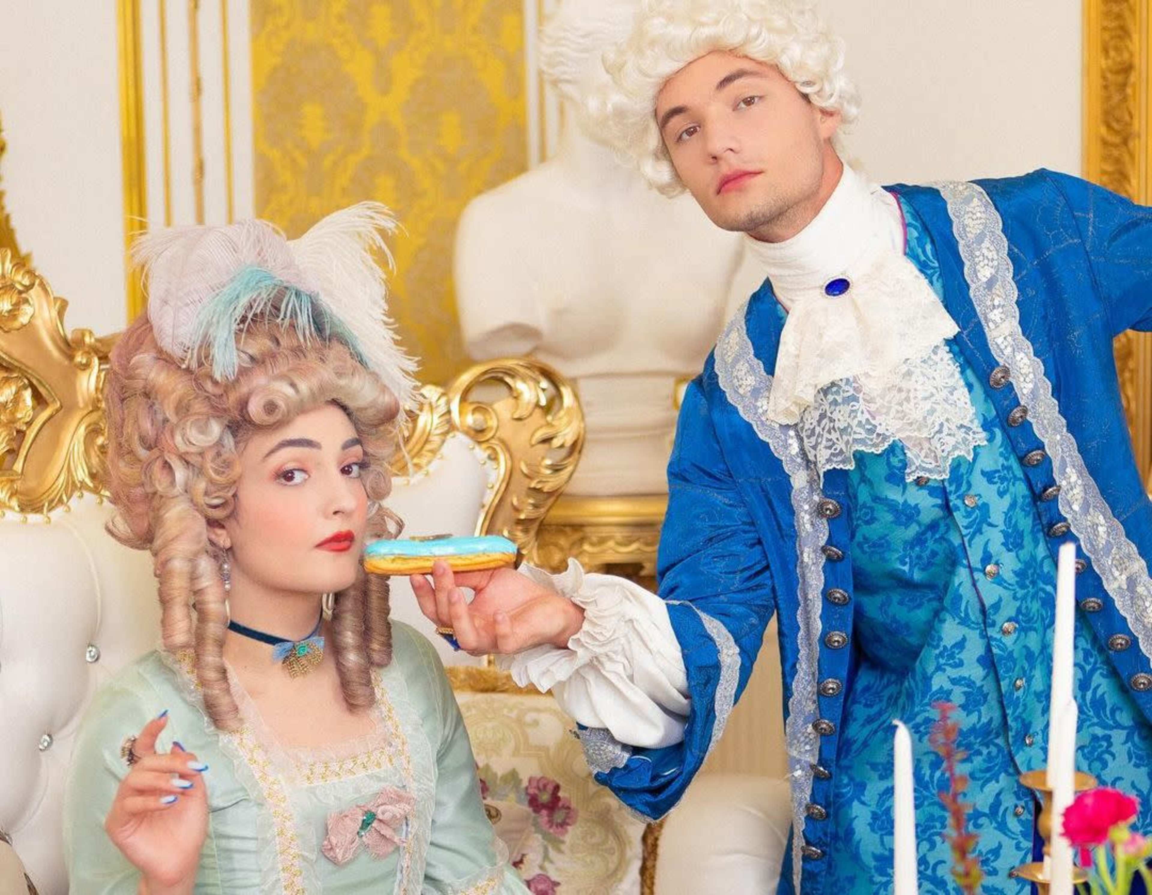 A man in an elaborate blue costume offers a macaroon to a woman in a pastel gown with a large wig, seated in an ornate room.