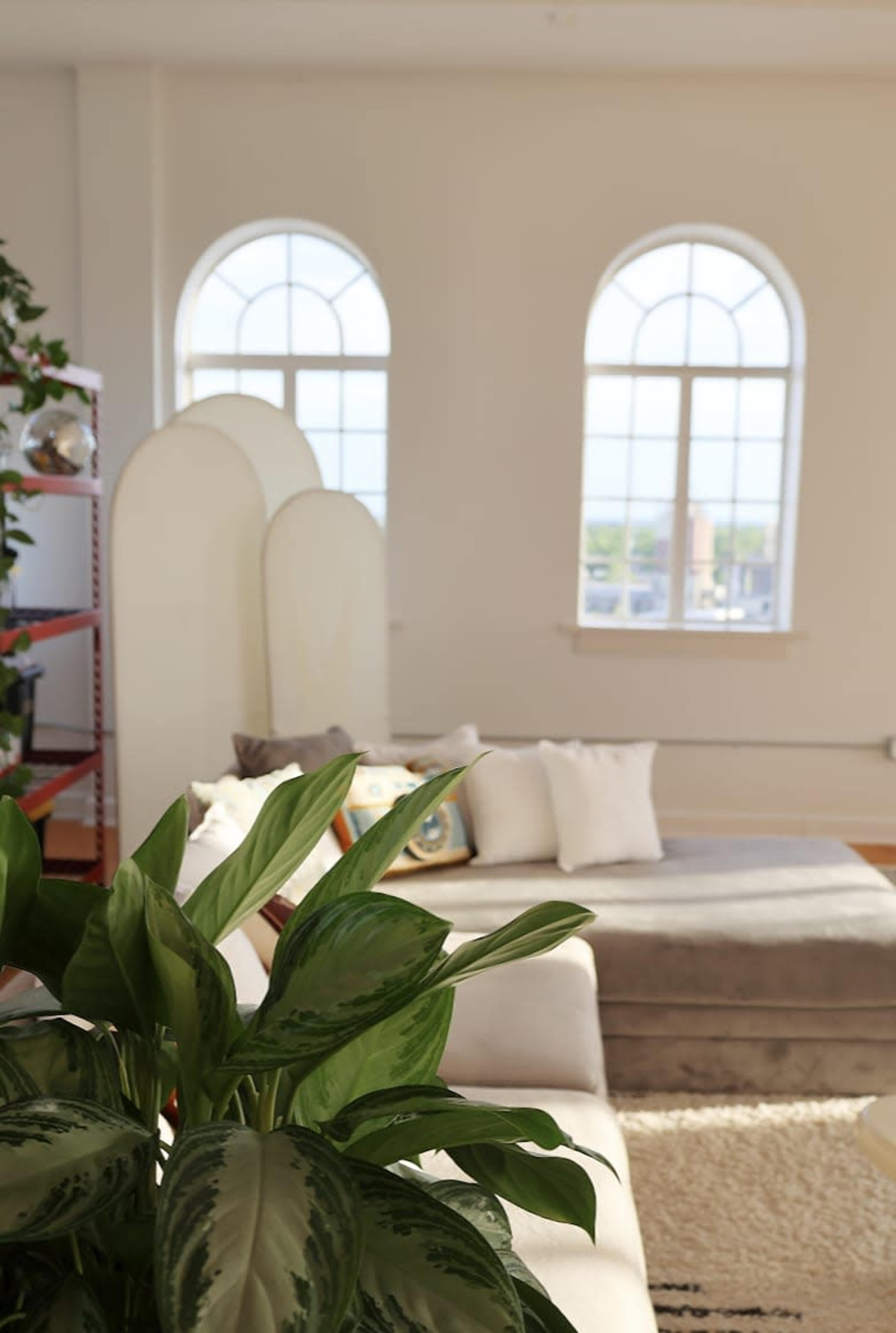 A sunlit room features a cream-colored sofa with throw pillows, a decorative plant, and arched windows overlooking a cityscape.