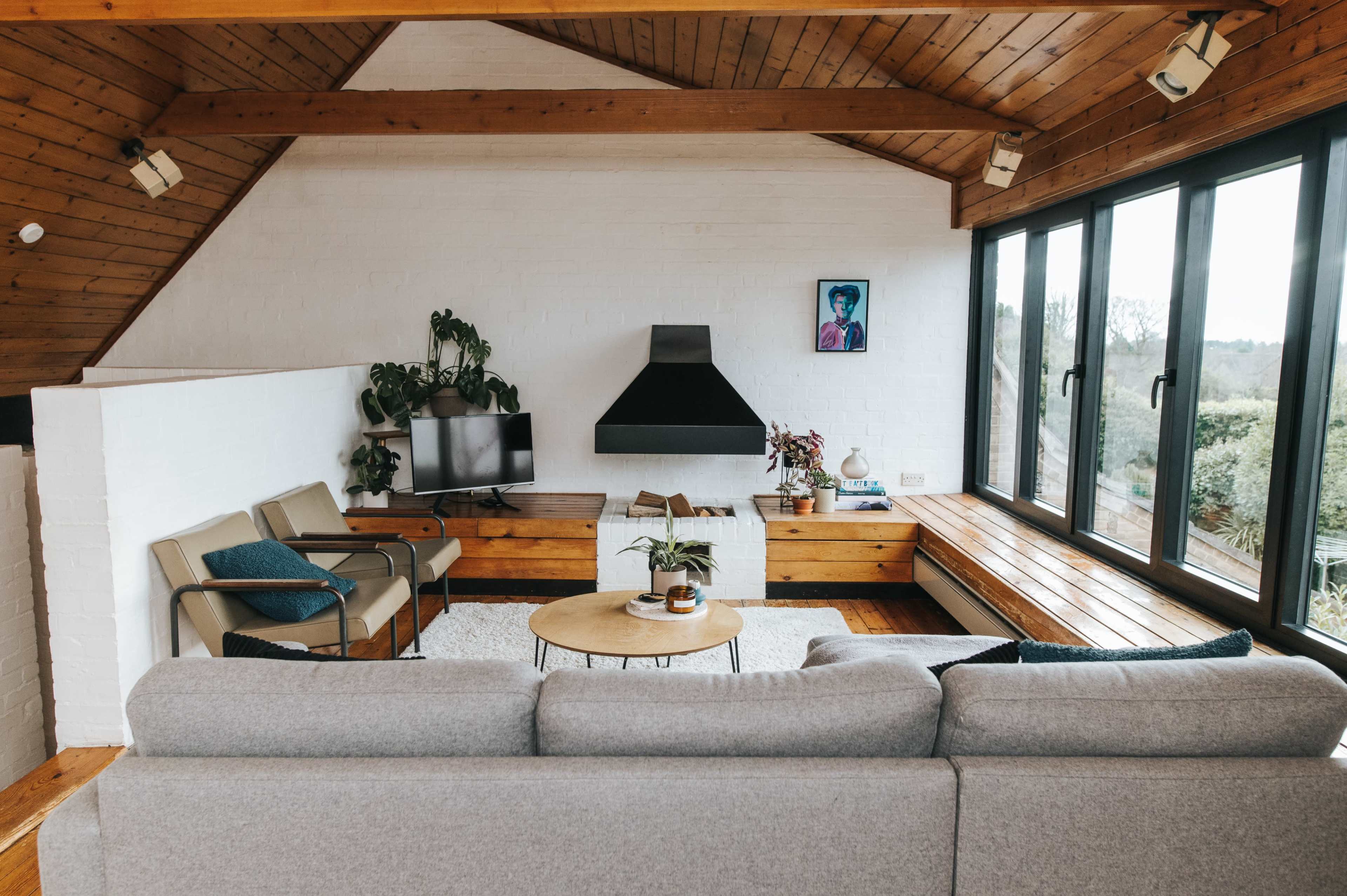 A modern living room features a gray sectional sofa, wooden accents, large windows, and a minimalist fireplace.