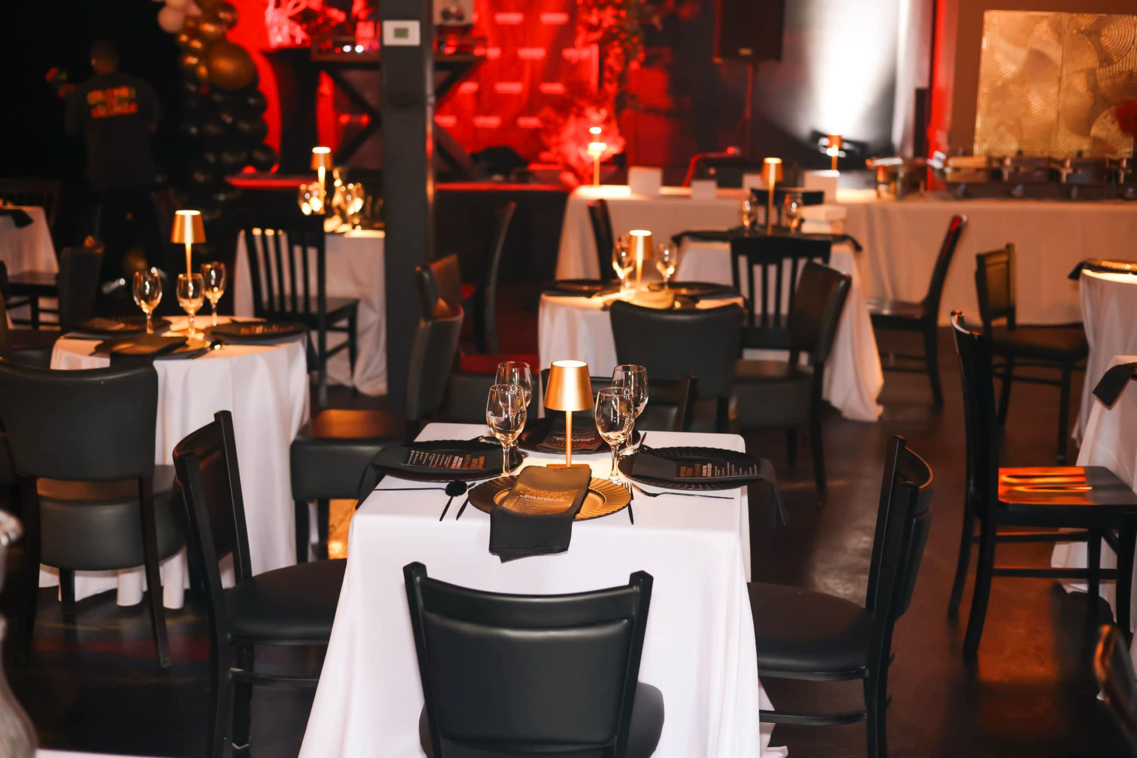 The image shows a dining area set up with black chairs and white tables, each adorned with glassware and small decorative lights, in an elegantly decorated venue.