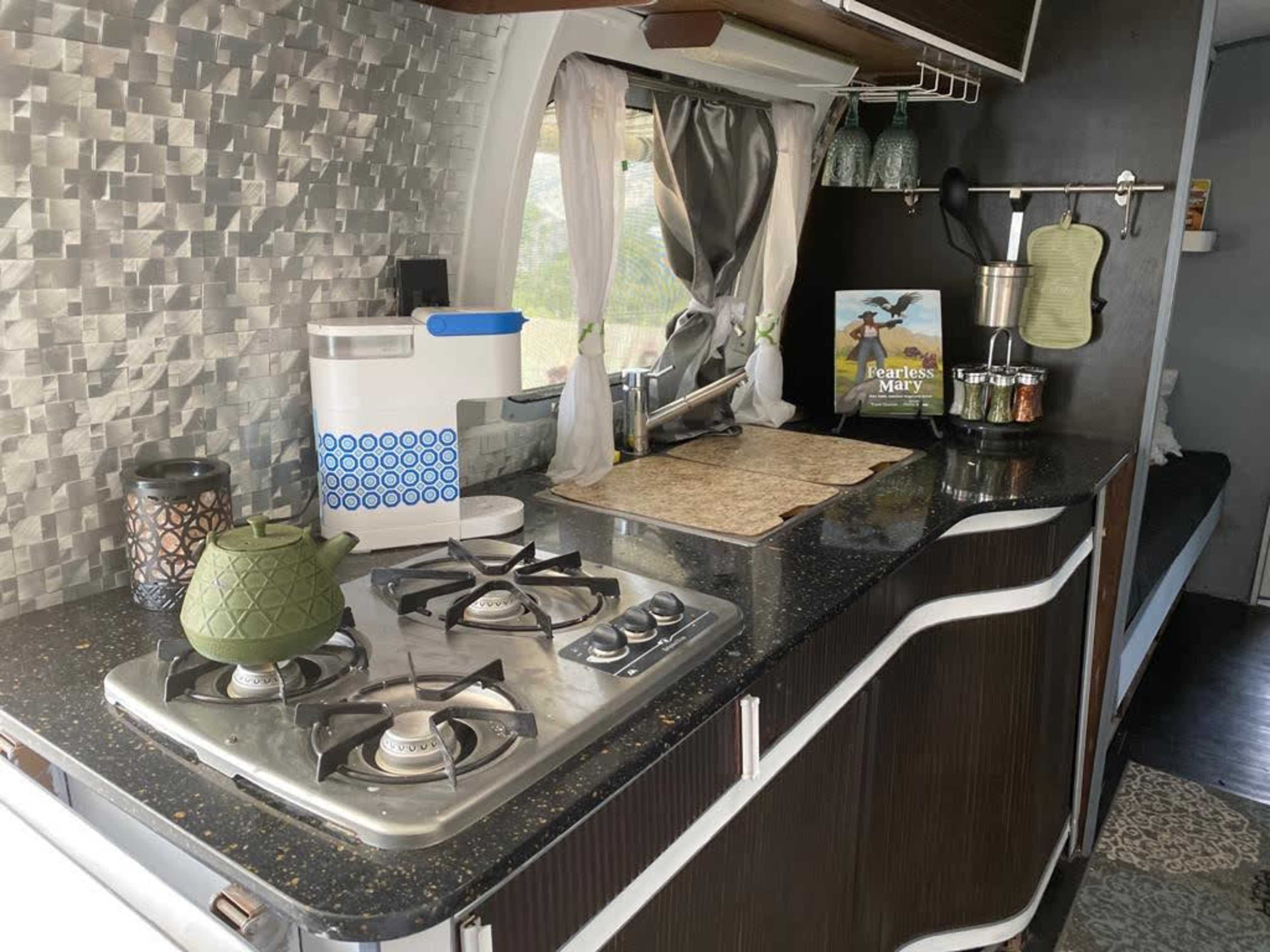 The image shows a modern kitchen with a gas stove, a green teapot, and various kitchen accessories on a countertop with a textured backsplash.