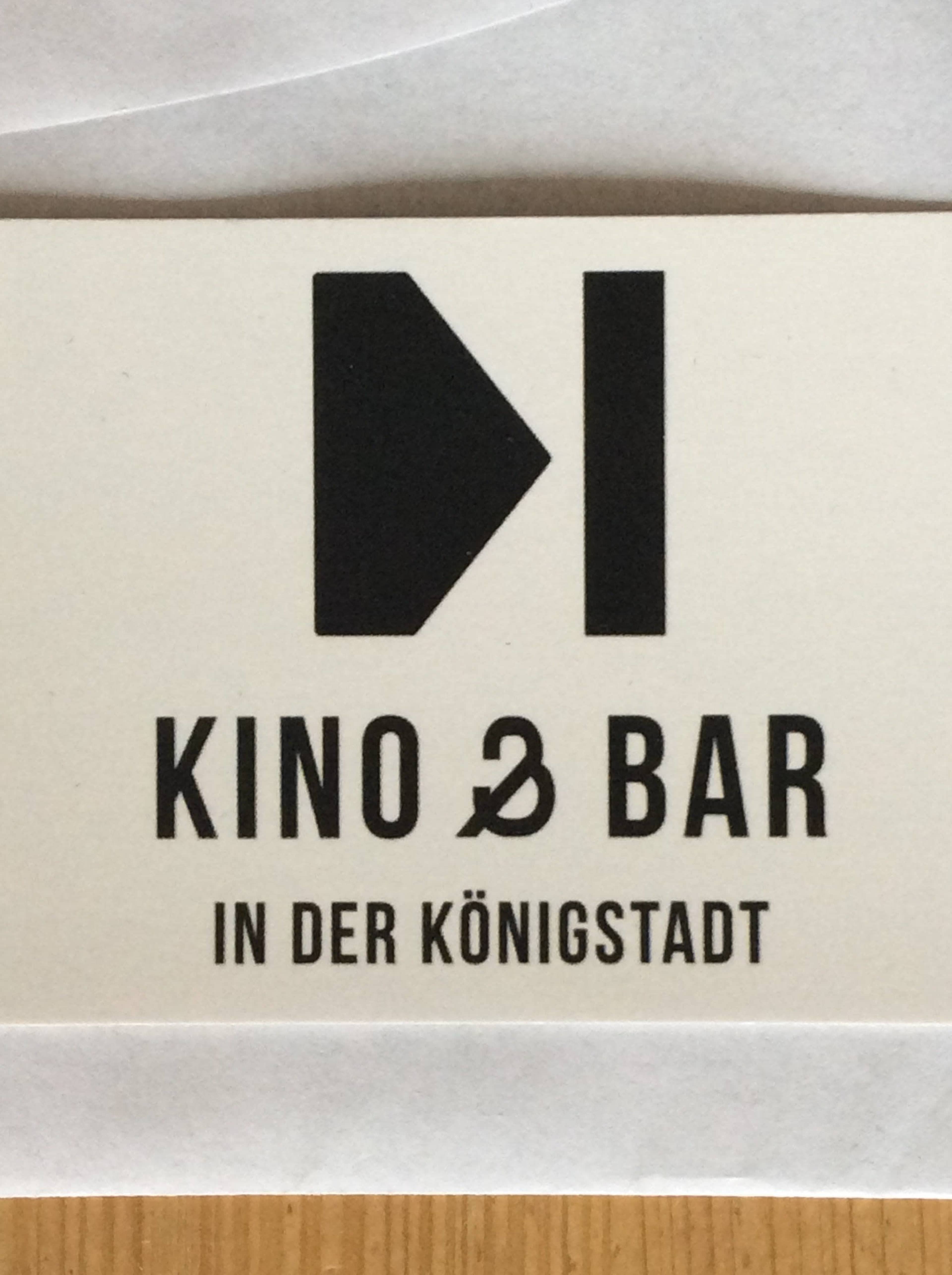 The image shows a business card with a logo featuring a stylized letter "D" and the text "KINO & BAR IN DER KÖNIGSTADT."