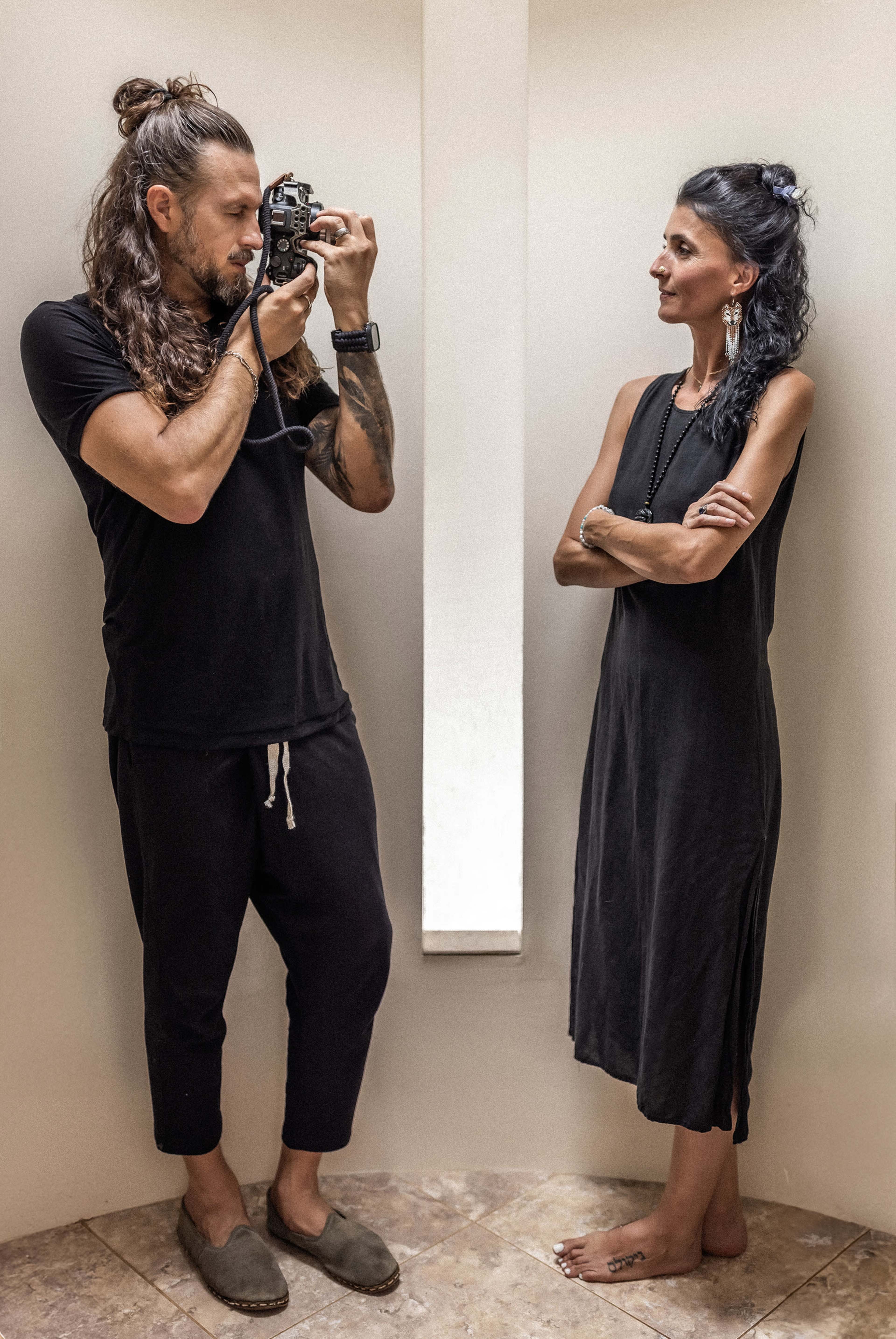 A man holding a camera stands opposite a woman with arms crossed, both dressed in black attire against a neutral wall.
