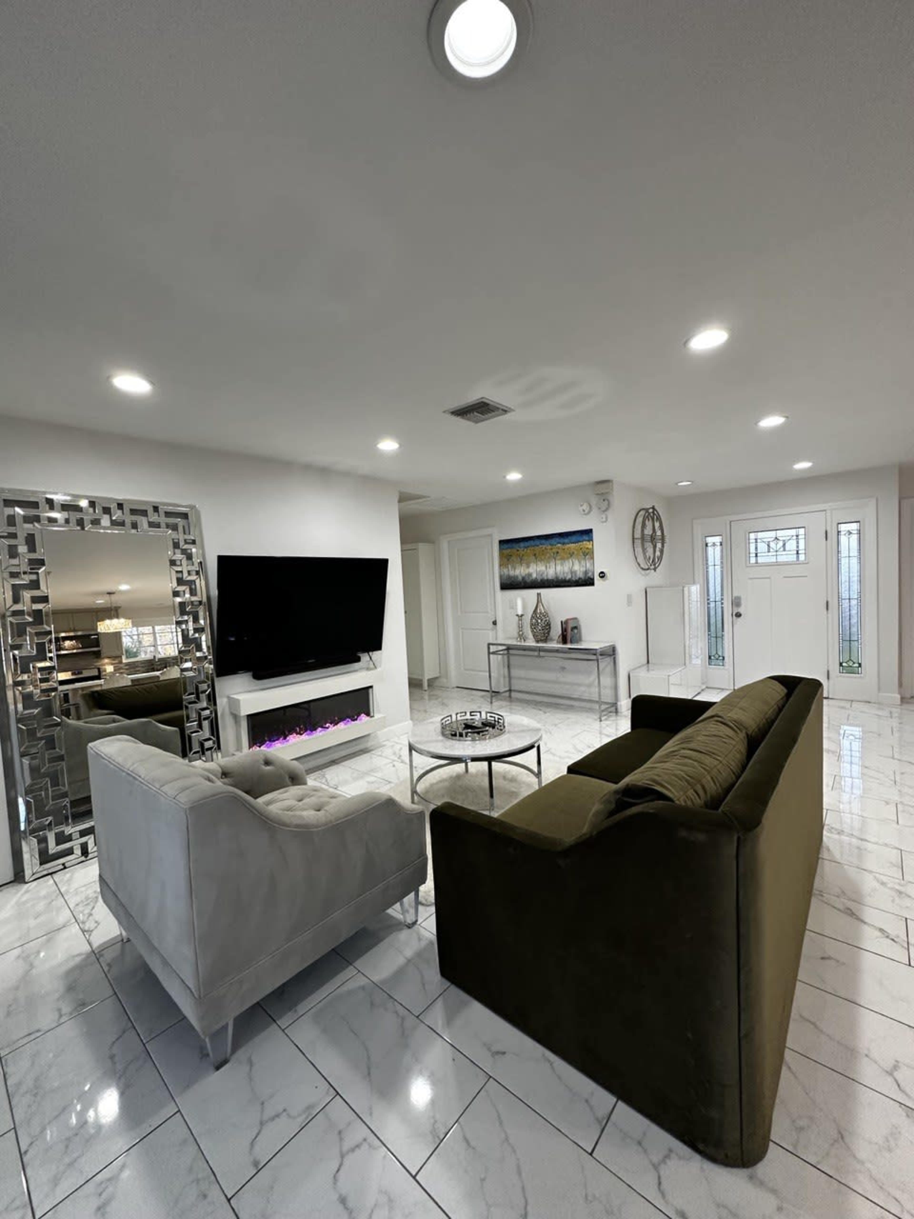 The living room features two couches facing a wall-mounted television, with a coffee table in the center and a large mirror reflecting the space.