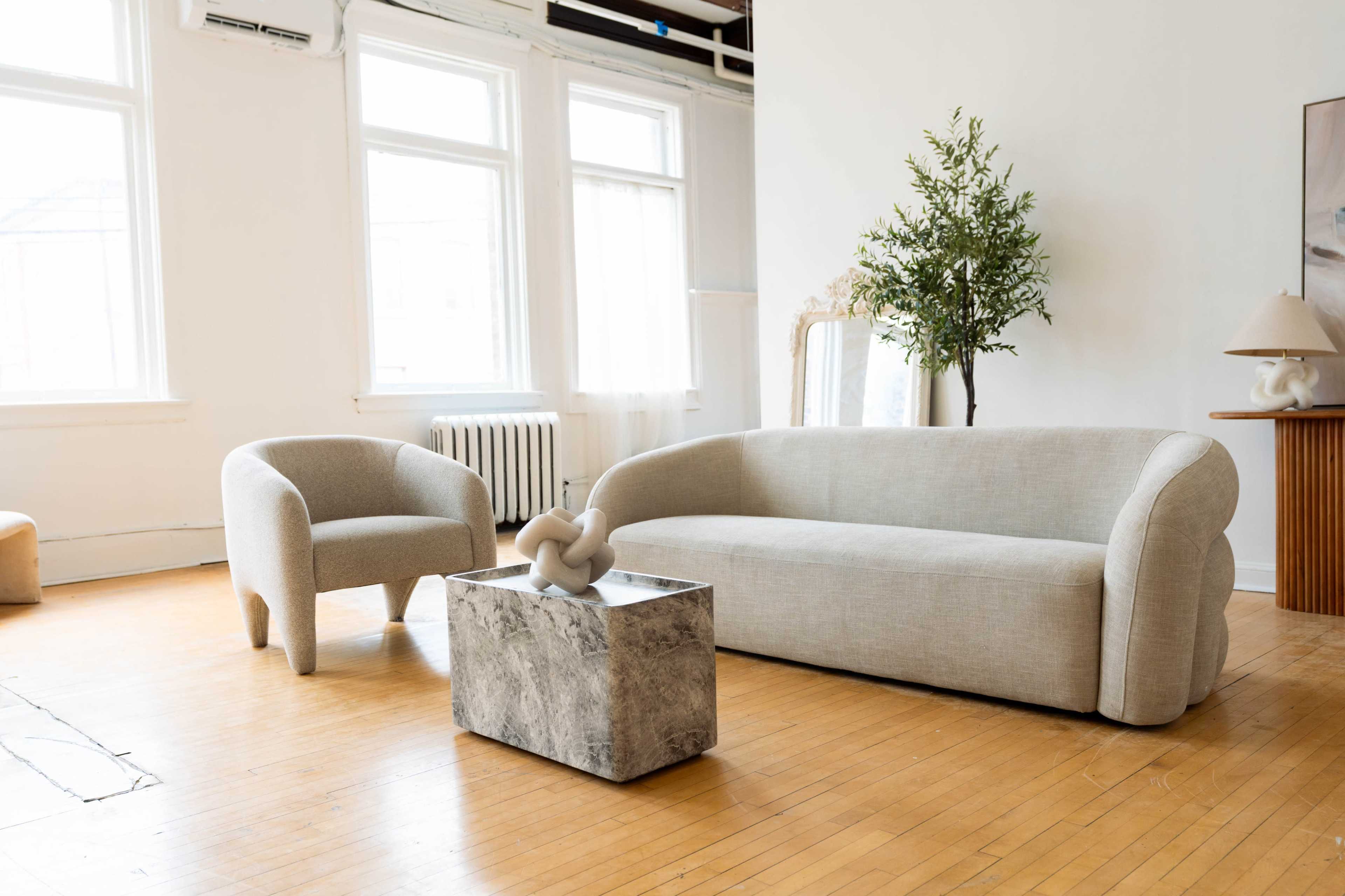The image features a minimalist living room with a light gray sofa, a matching armchair, a marble coffee table, and a potted plant next to large windows.