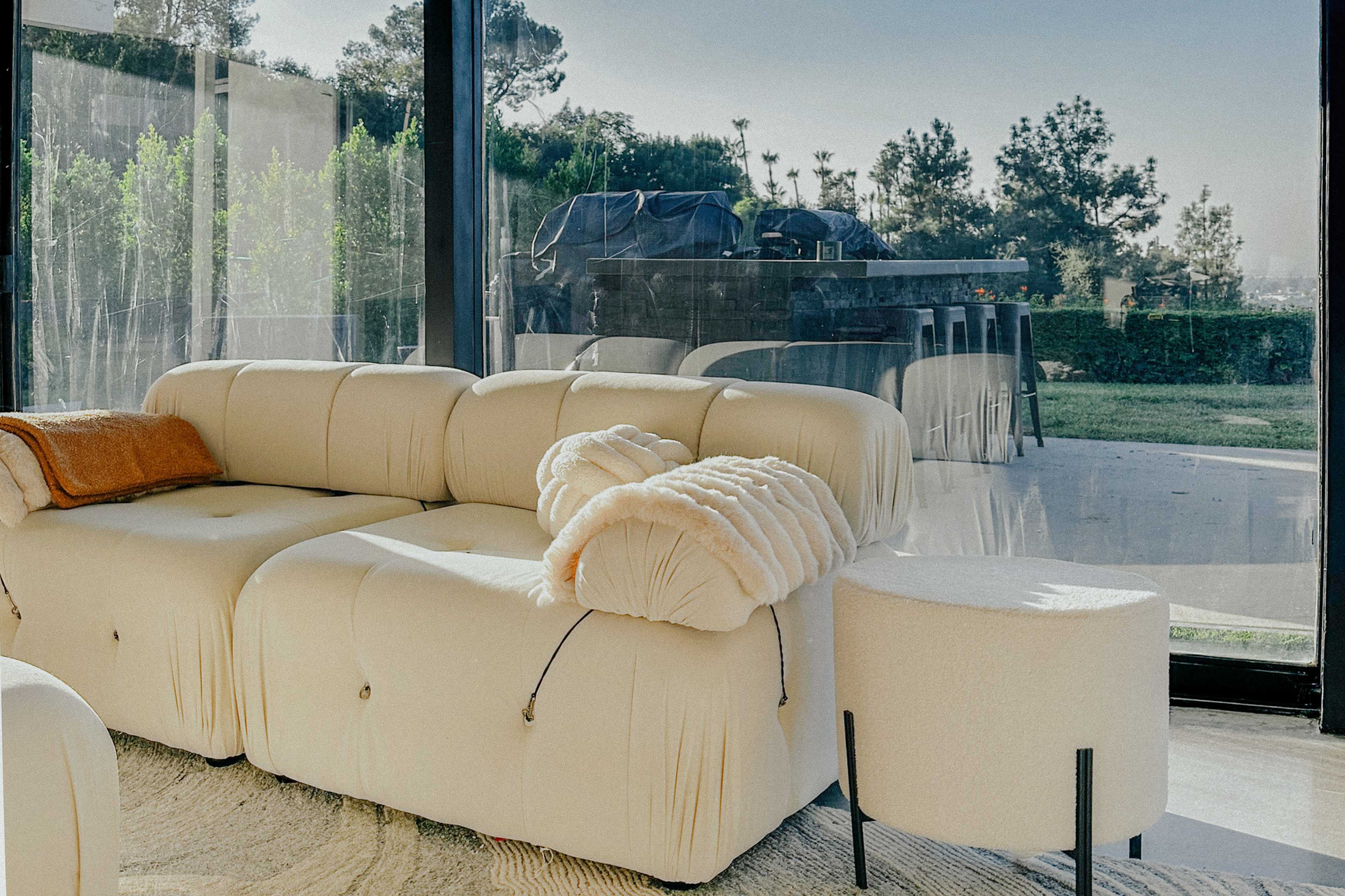 A modern living room with a cream-colored sofa and a small round table, while a grill is visible outside through large glass windows.