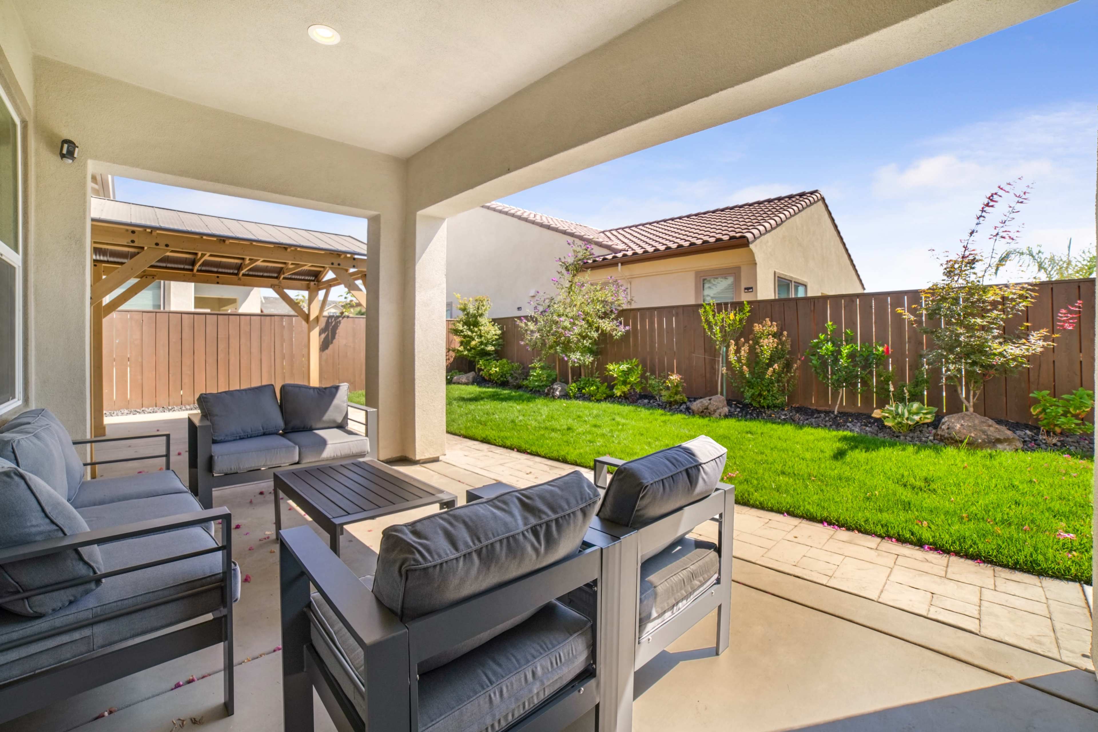 A covered patio features gray outdoor seating and a table, overlooking a green lawn and flower beds with a fence in the background.