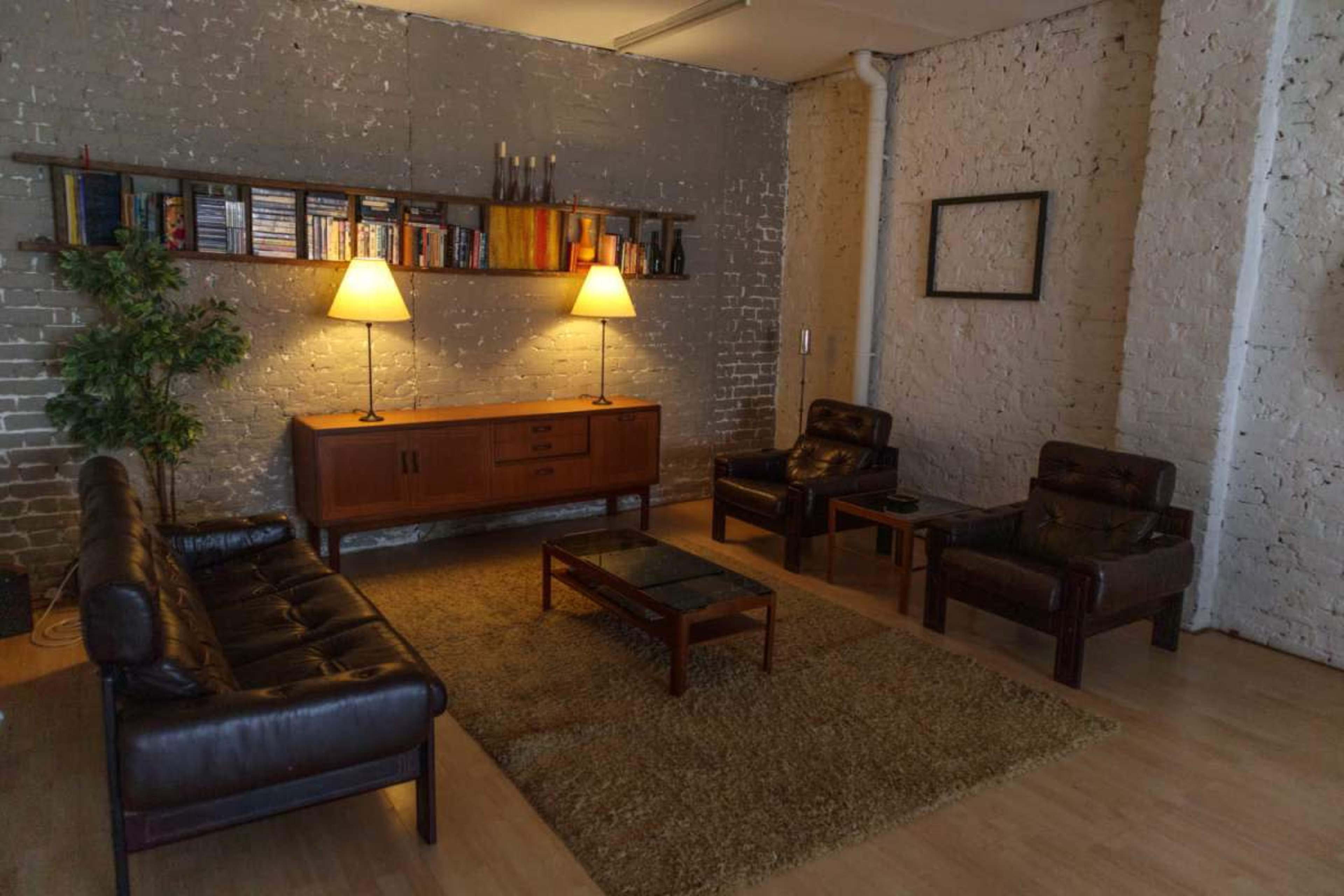 The image depicts a modern living room with a seating area featuring two leather sofas, two armchairs, a coffee table, and a shelf with books against a textured gray wall.