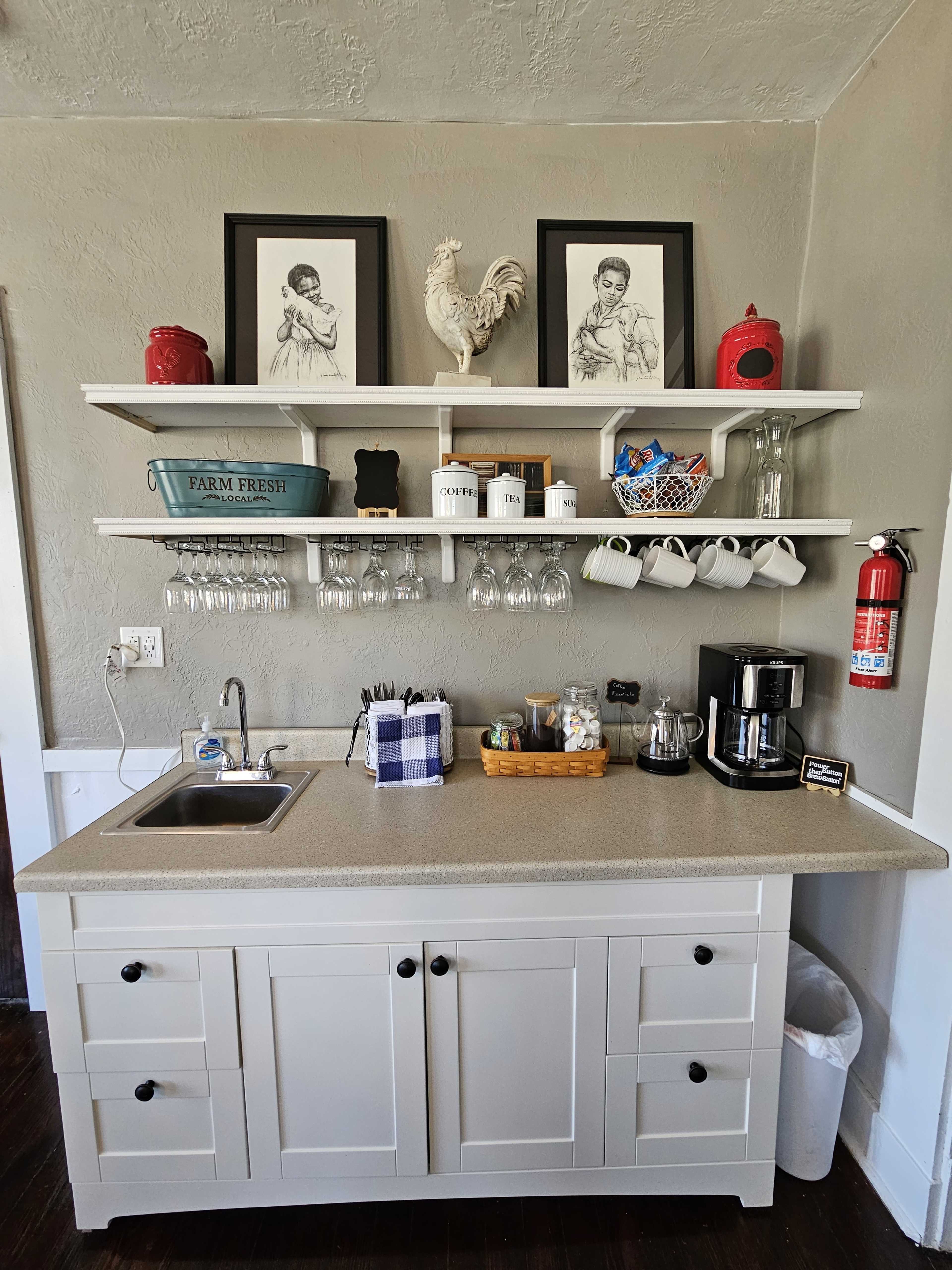A simple kitchen area featuring a countertop with a sink, coffee maker, and storage for mugs, alongside a shelf displaying framed artwork and a rooster statue.