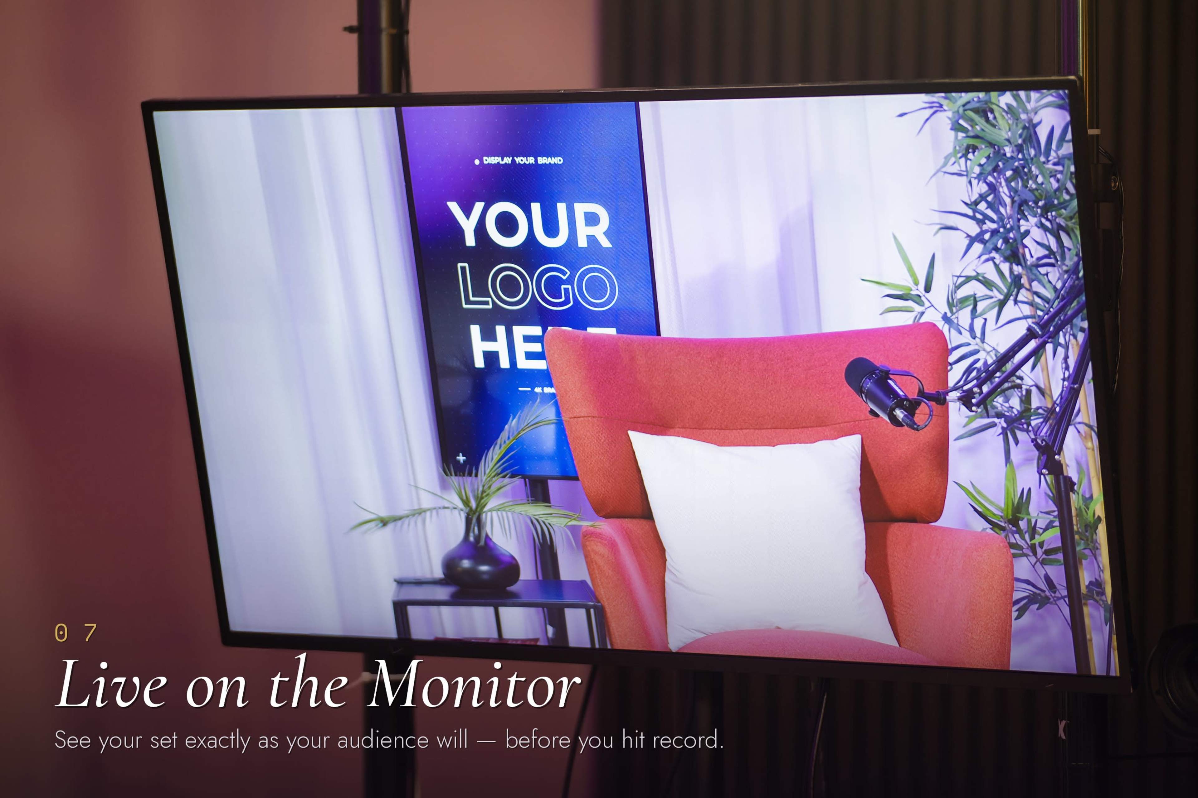 The image shows a large monitor displaying a set with a red armchair, a microphone, and a backdrop featuring a placeholder for a logo.