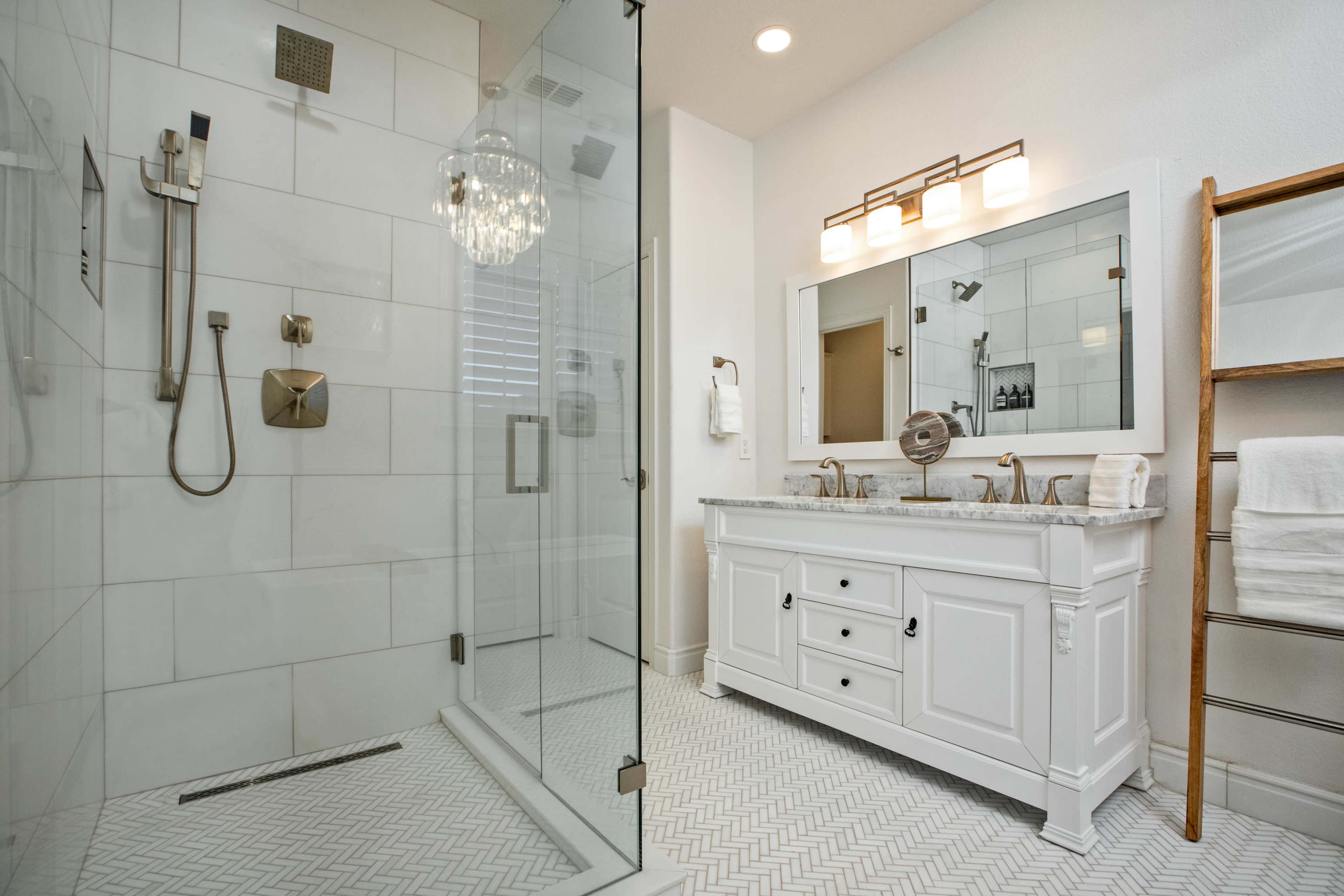 The bathroom features a glass-enclosed walk-in shower, a double vanity with a marble countertop, and a large mirror, all set against white tiled walls and a patterned floor.