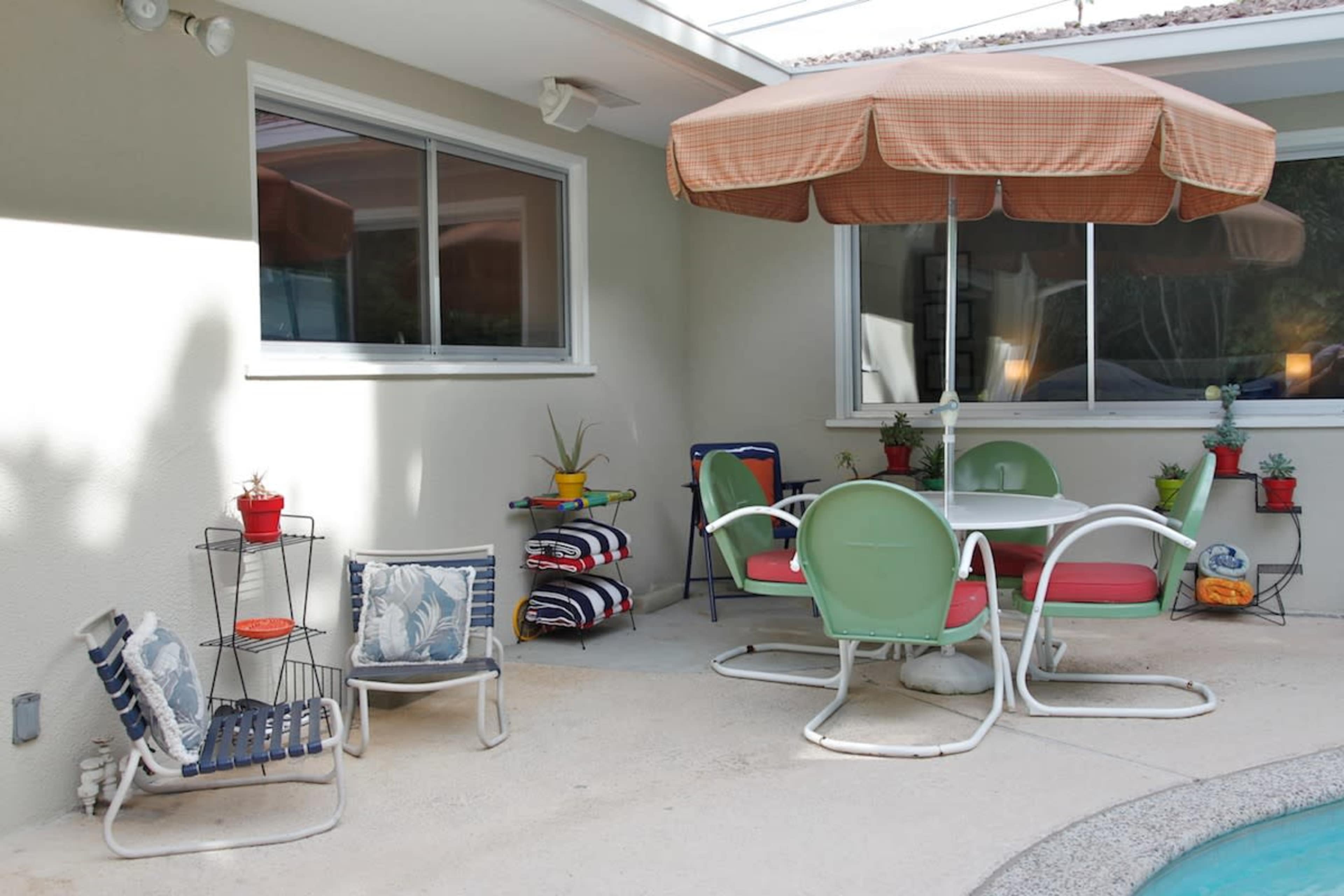 A patio area features a round table with chairs and an umbrella, surrounded by planters and lounge chairs.