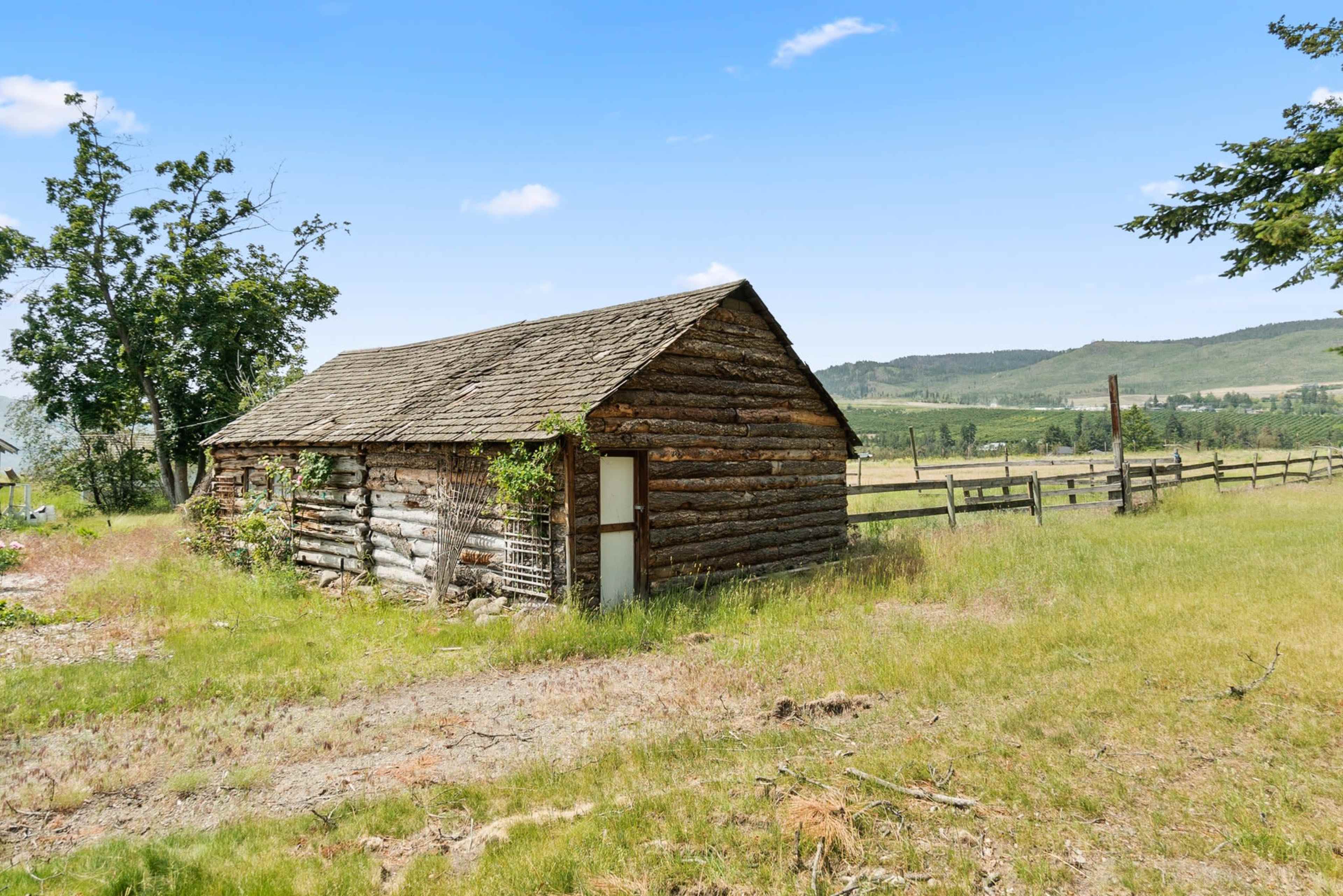 A weathered log cabin stands in a grassy field surrounded by trees and hills in the background.