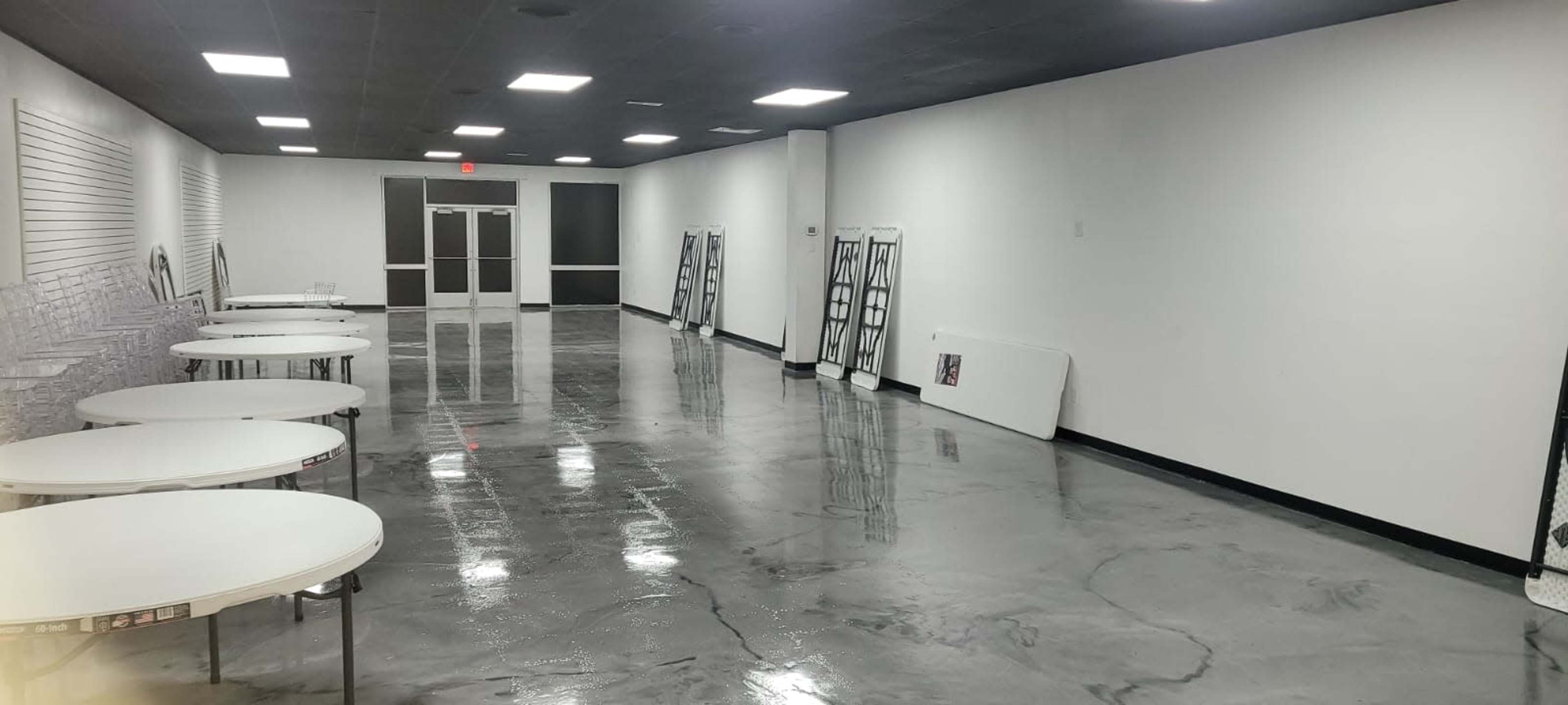 The image shows a large, empty room with polished concrete floors and several round tables set up along the sides.