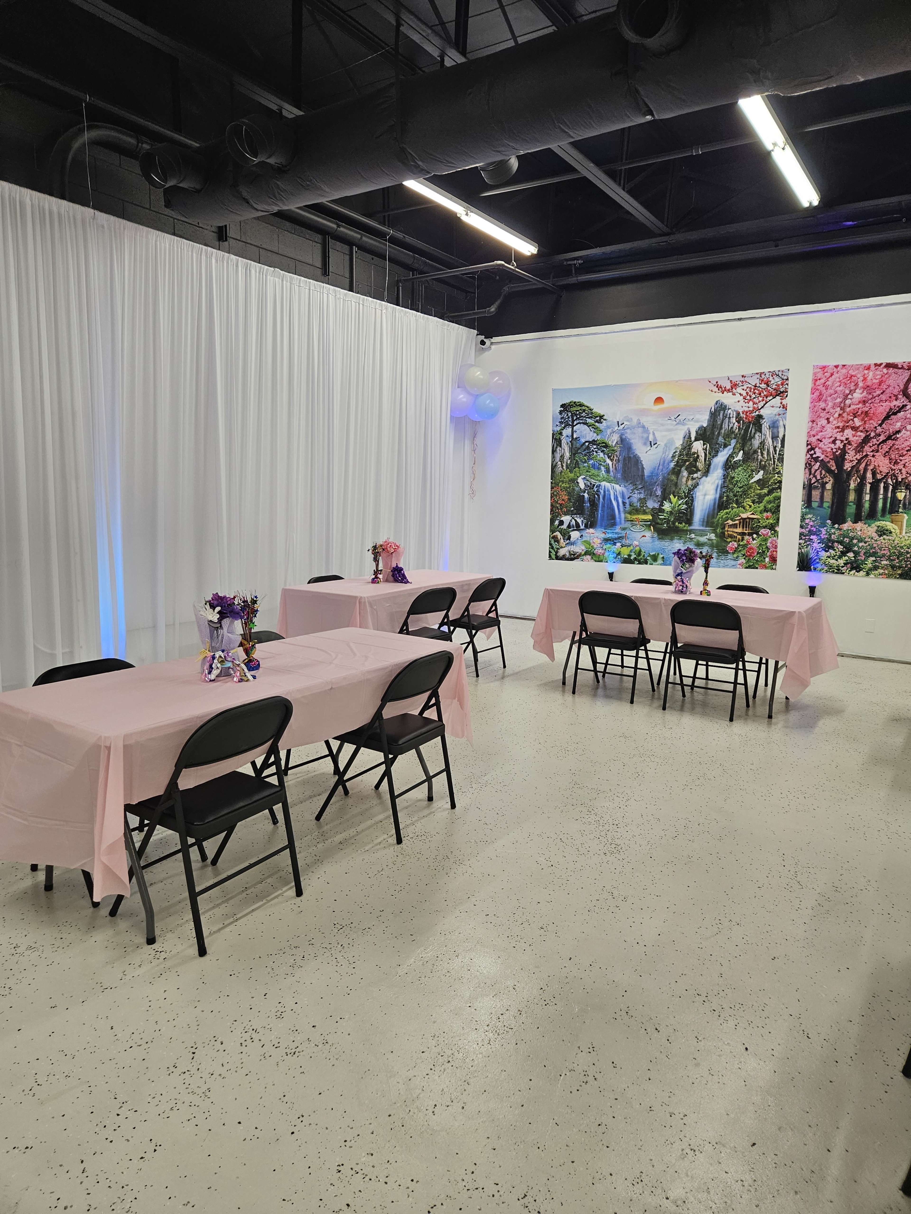 A party space with several tables covered in light pink tablecloths, black chairs, and decorative floral centerpieces, set against a backdrop of large nature-themed wall murals.