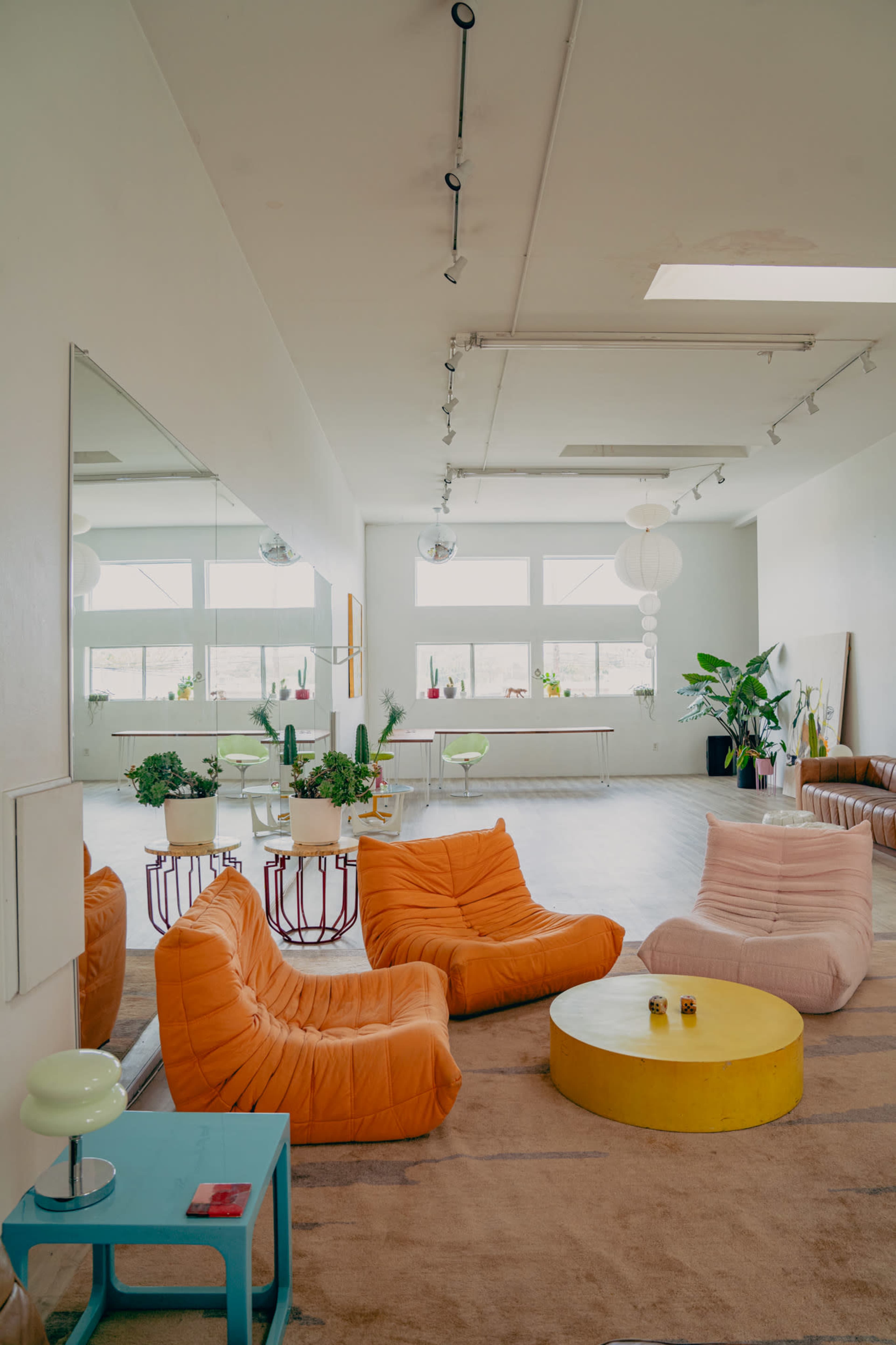 The scene features a spacious, brightly lit interior with colorful seating, a round coffee table, and numerous potted plants near large windows.