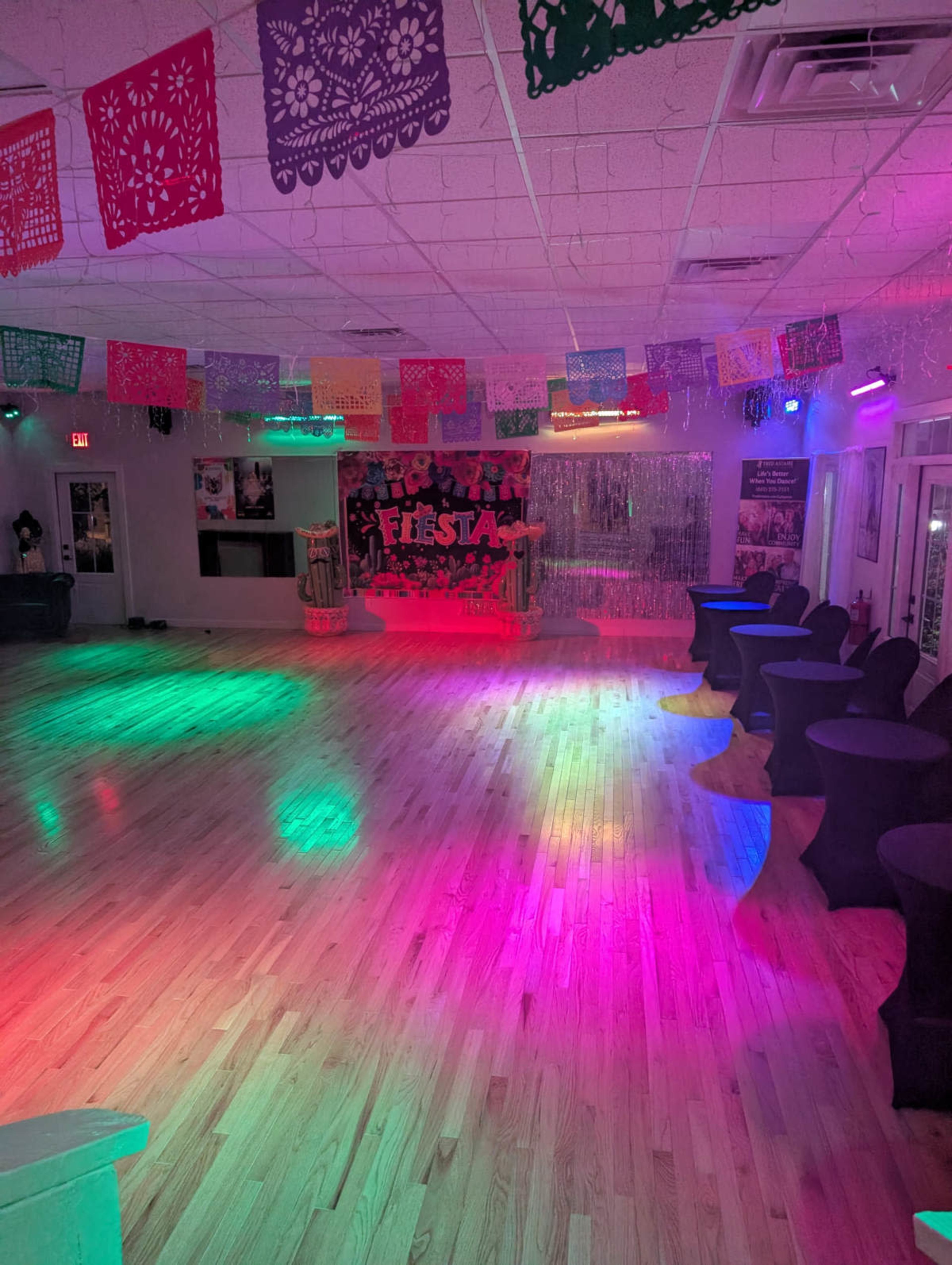 The image shows a brightly lit event space decorated with colorful papel picado banners and spotlighting, featuring black table covers arranged around the floor.