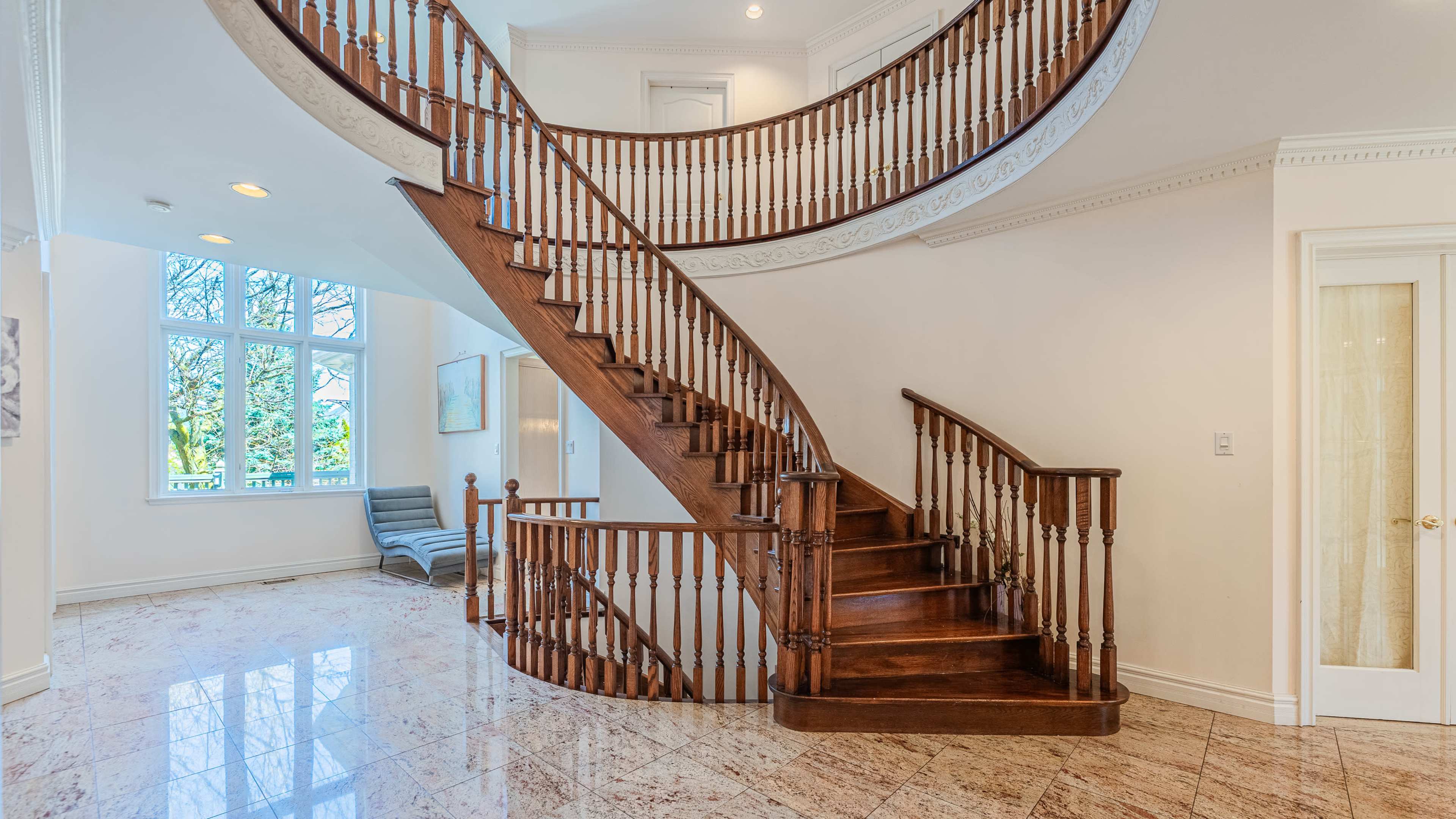 The image shows a curved wooden staircase in a well-lit foyer with marble flooring and large windows.