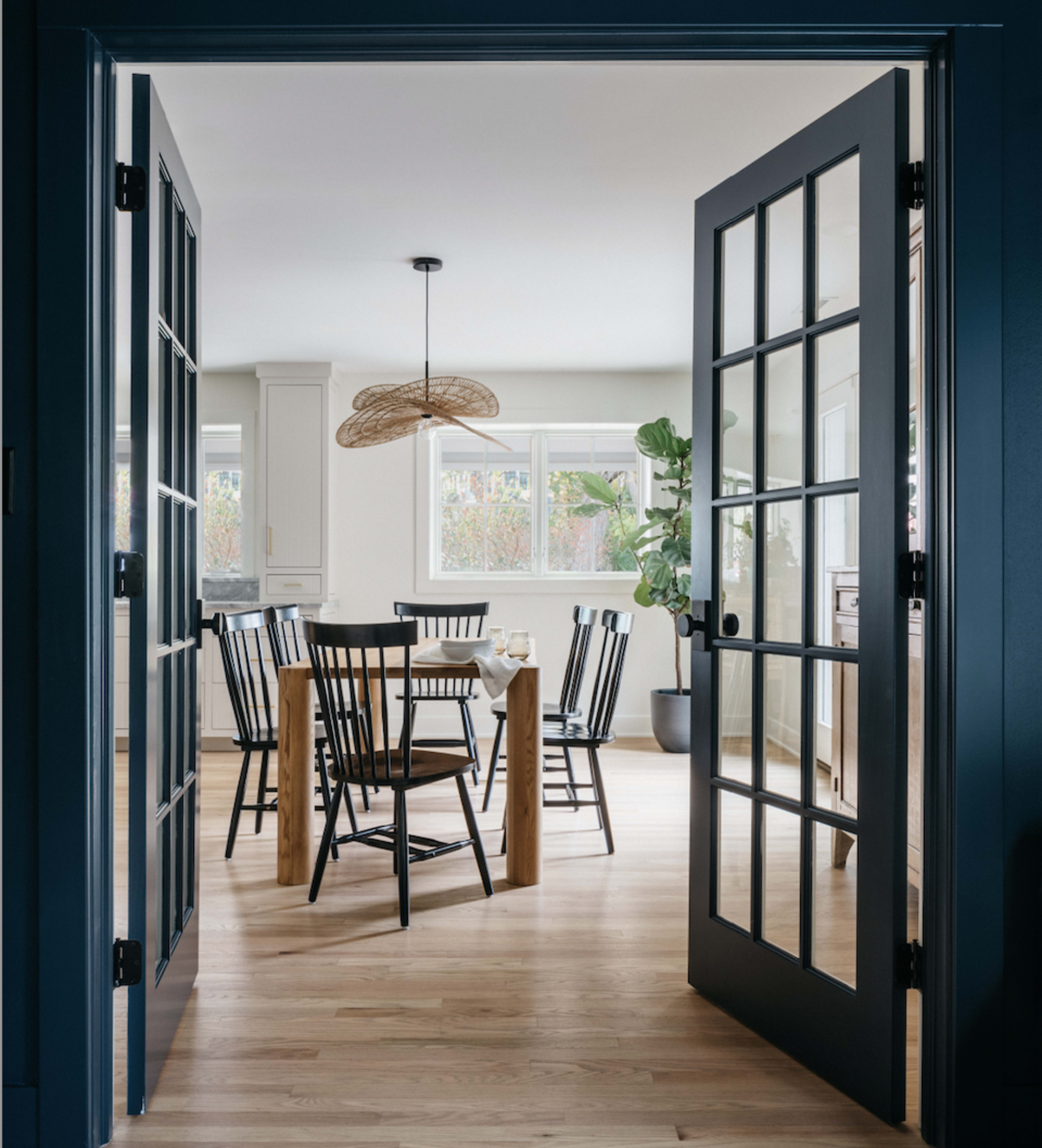 A dining area features a round table surrounded by black chairs, with large windows allowing natural light and a plant in the corner.