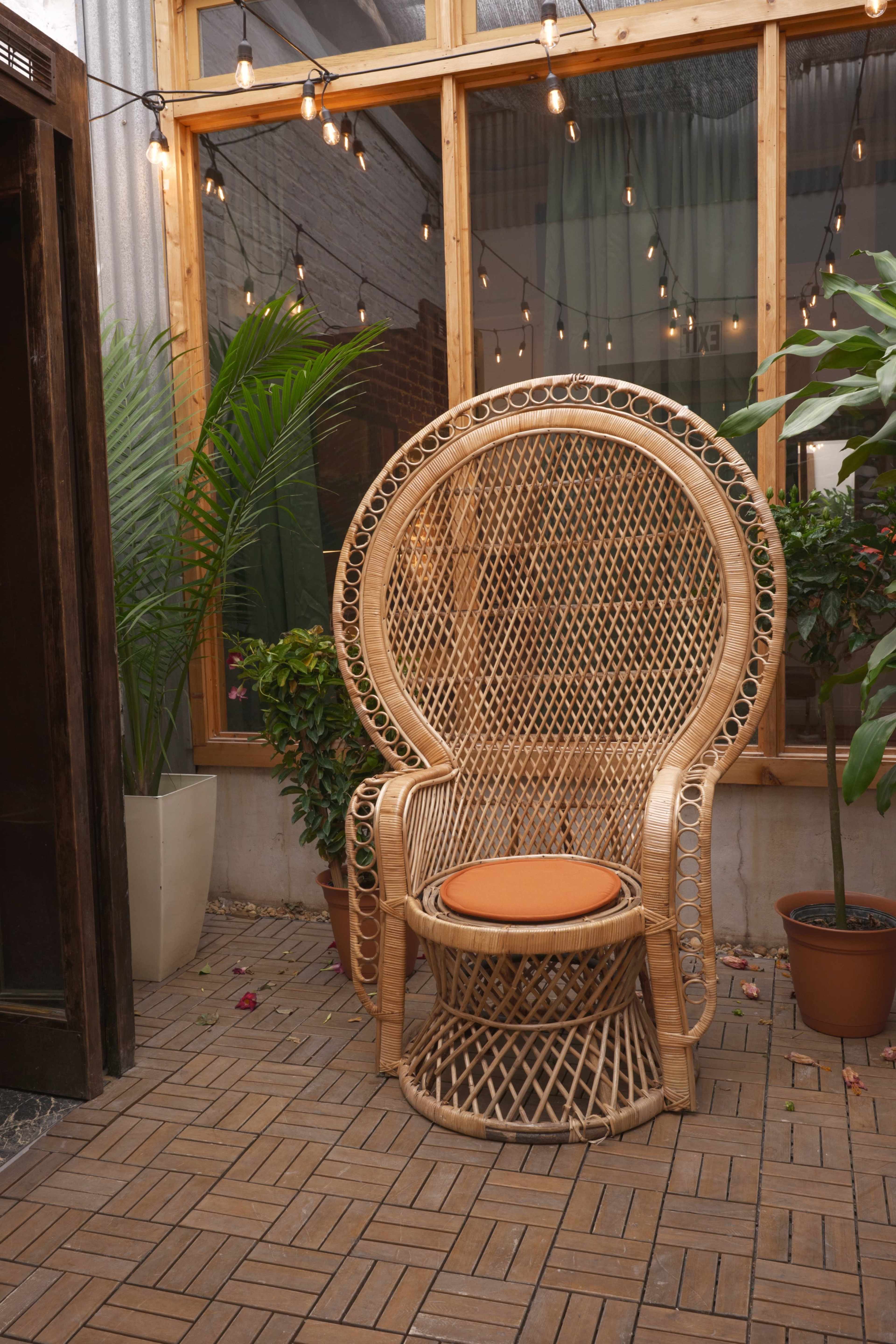 A large, intricately woven rattan chair with an orange cushion is positioned in a brightly lit interior space surrounded by greenery and string lights.