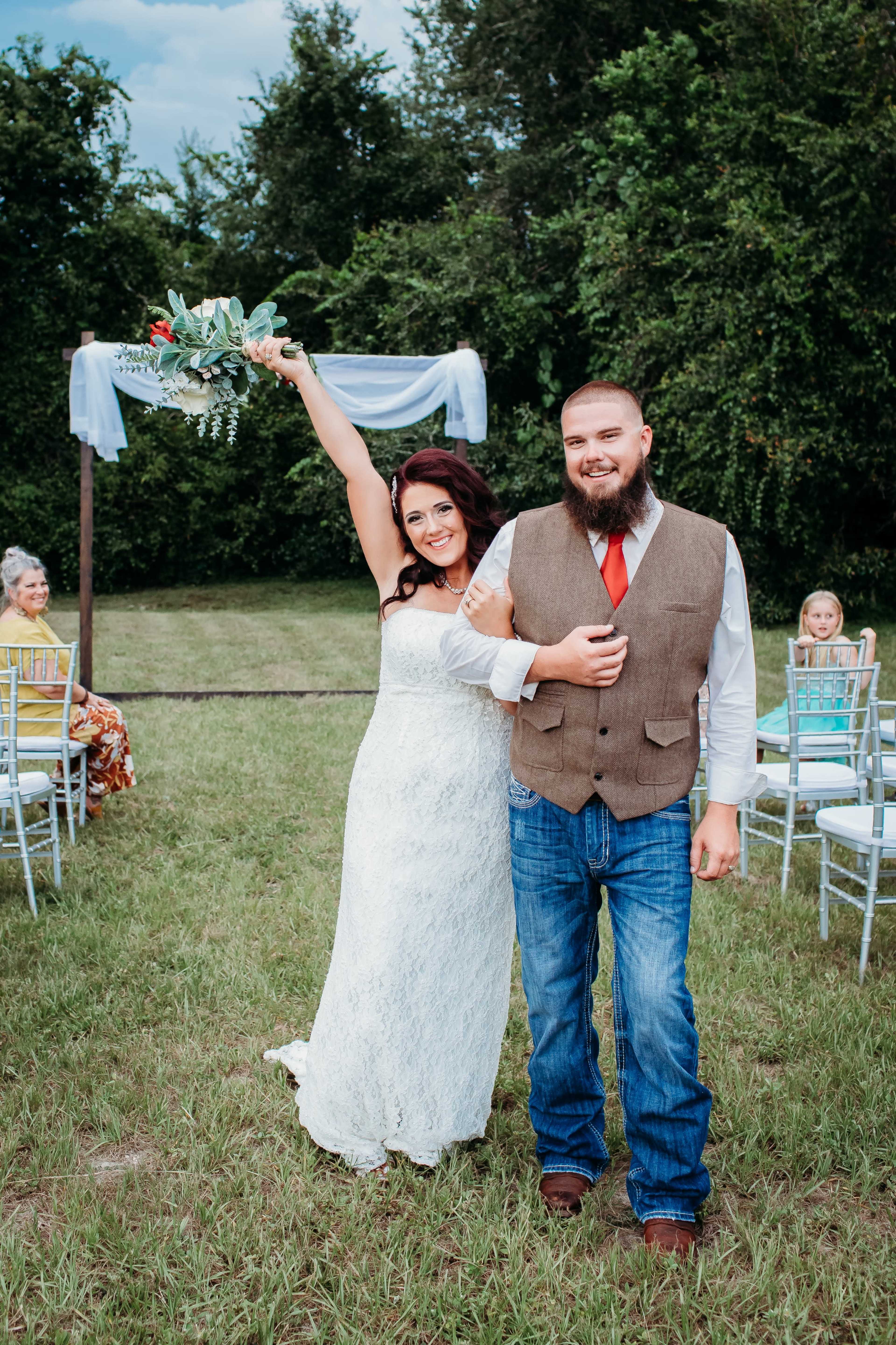 A bride in a white lace dress holds a bouquet aloft next to a groom in a vest and jeans, standing in an outdoor wedding setting with guests seated nearby.