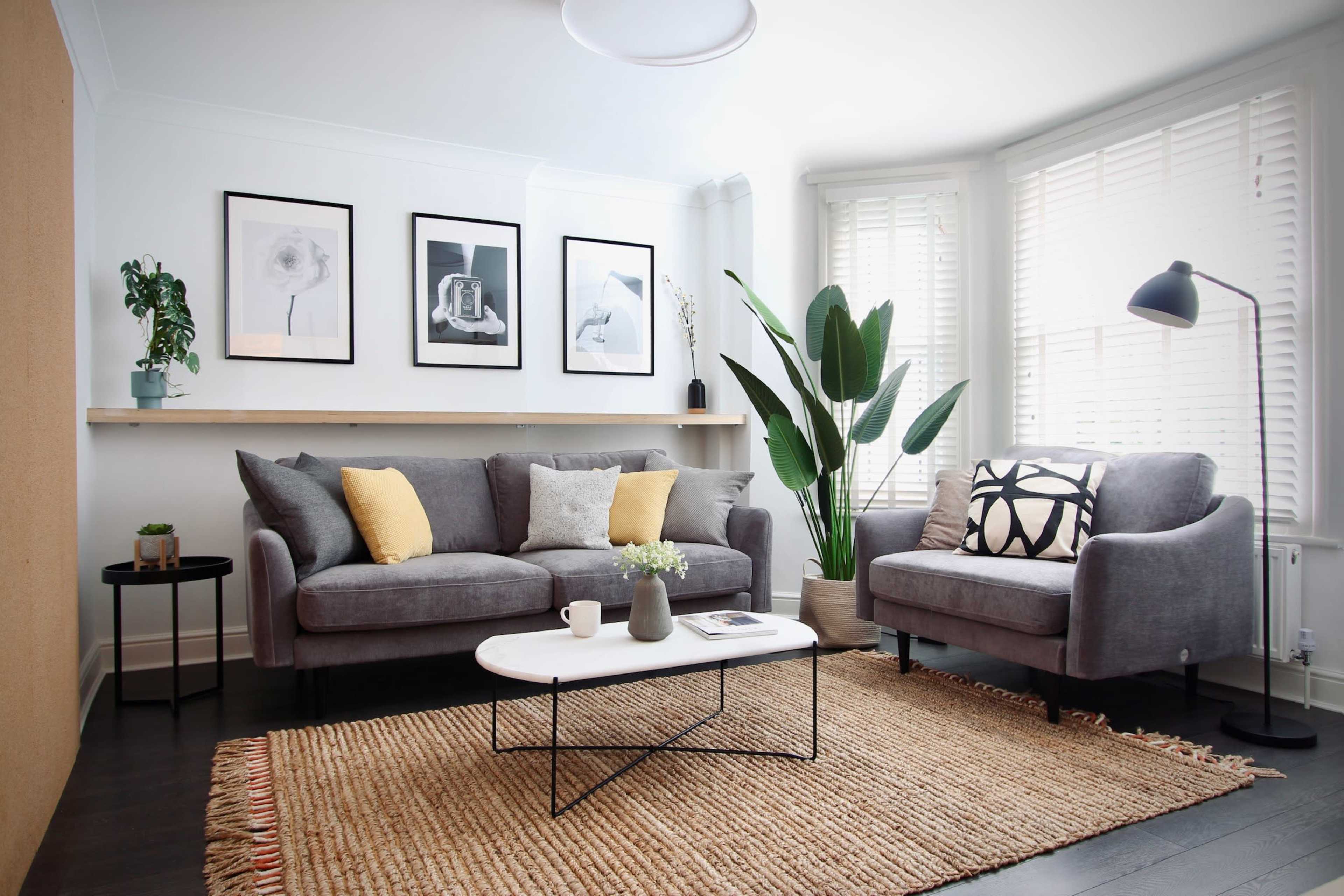 The image displays a modern living room with a gray sofa, white coffee table, and framed artwork on the walls, complemented by indoor plants and a natural fiber rug.